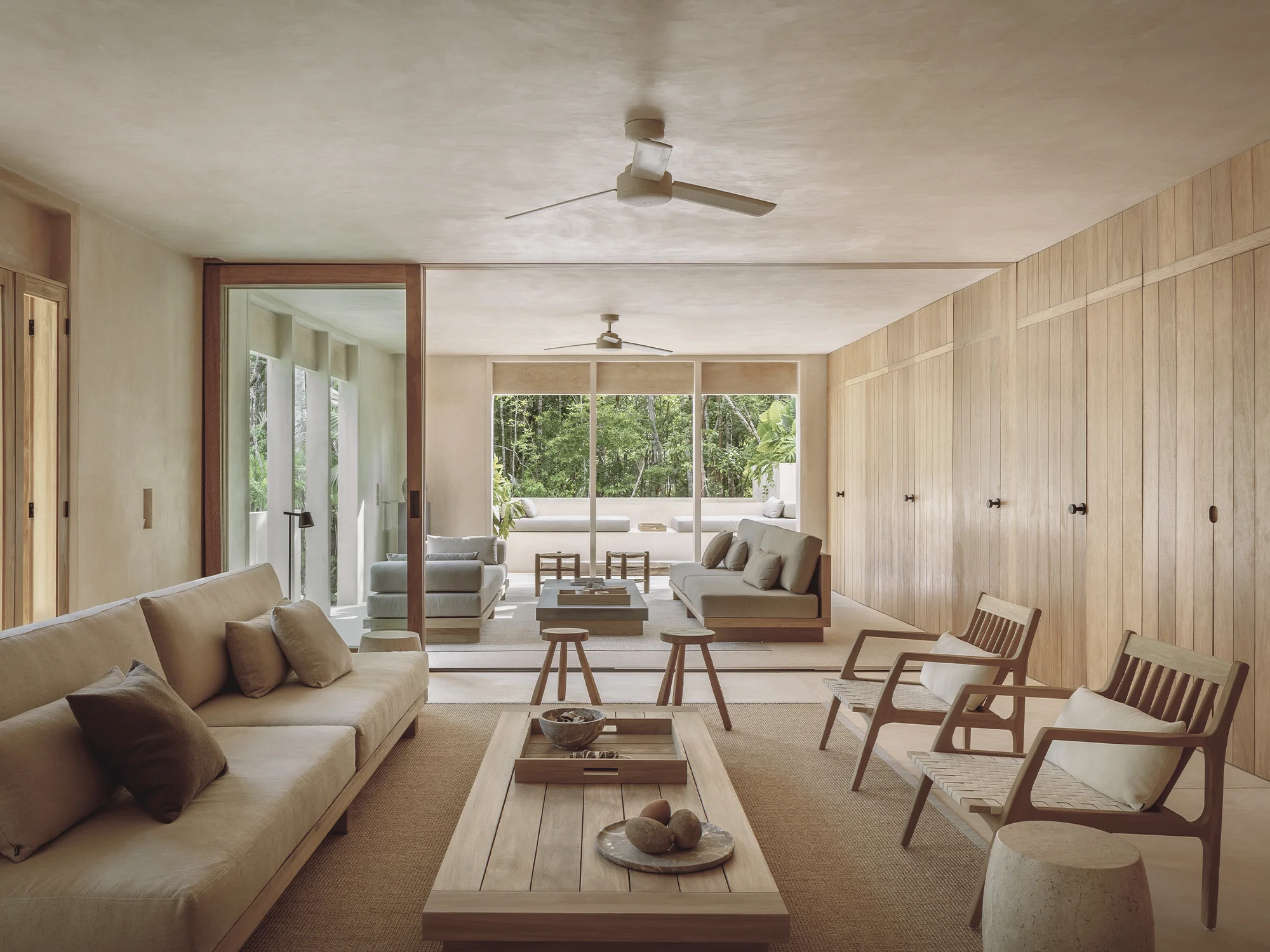 Modern living room with beige sofas, wooden chairs, coffee table, and large windows overlooking greenery.