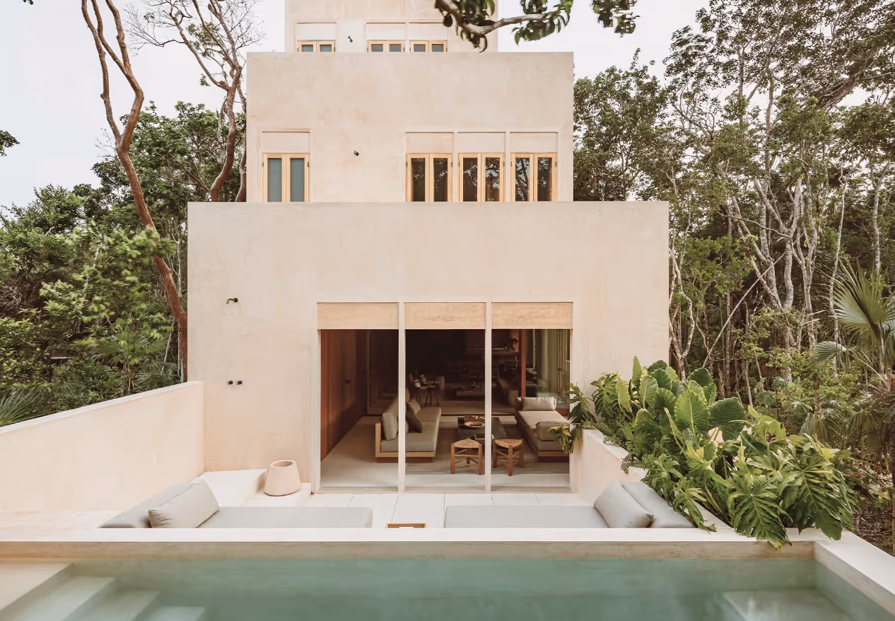 Modern beige house with large sliding glass doors opening to an outdoor seating area and a pool surrounded by trees and greenery.