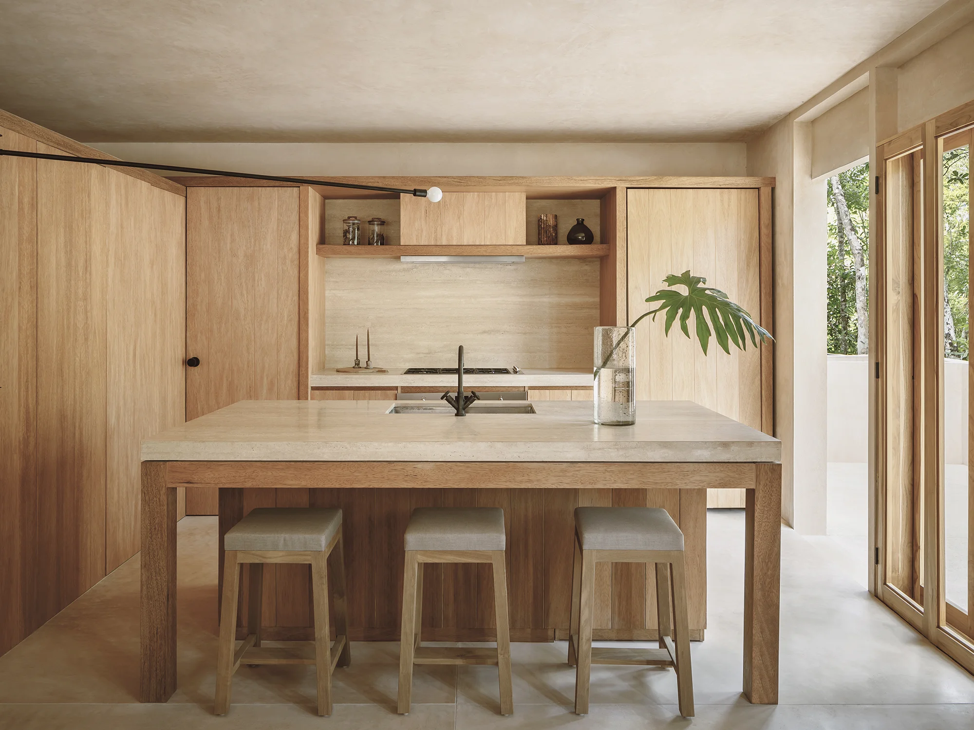 Minimalist kitchen with wooden cabinetry, beige stone countertop island, three wooden stools, and a large leaf in a glass vase.