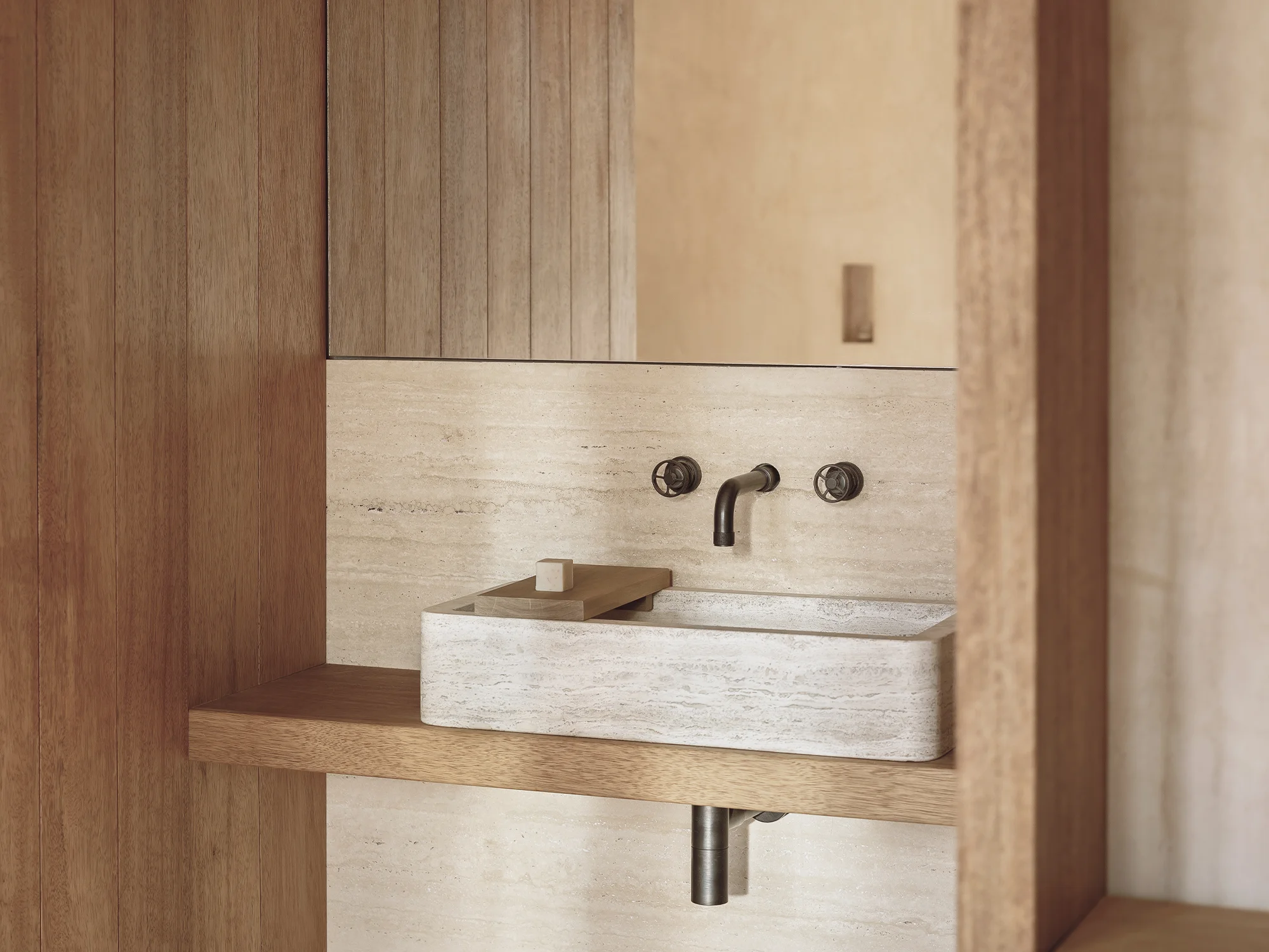 Modern bathroom sink with matte metal wall-mounted faucet and wooden shelf, set against beige stone and wood panel walls.