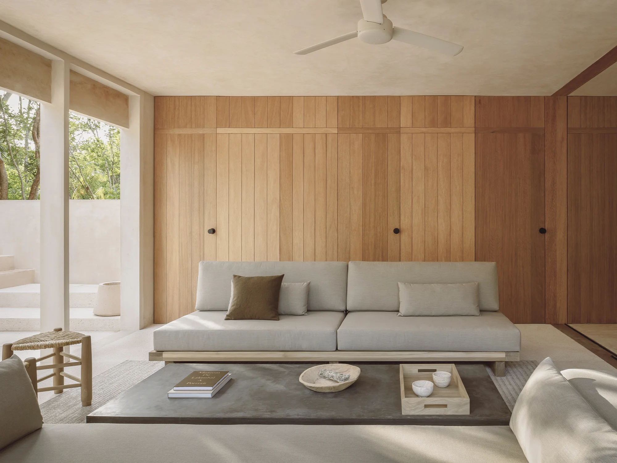 Minimalist living room with light gray sofas, wooden slatted wall, concrete coffee table with decor, and large open windows showing greenery outside.