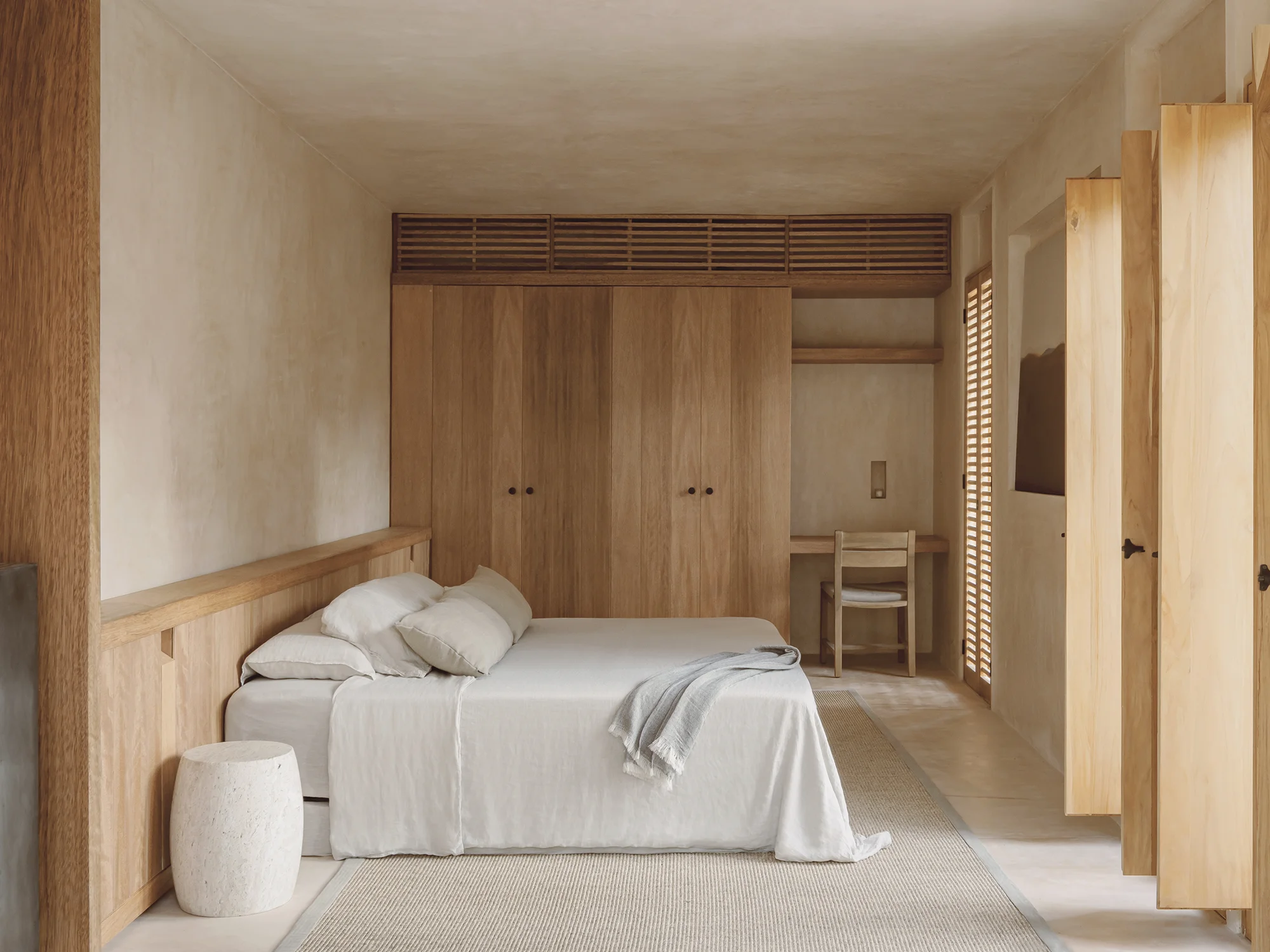 Minimalist bedroom with beige walls, wooden wardrobe, bed with white linens and pillows, small white stool, wooden desk and chair by window with wooden shutters.
