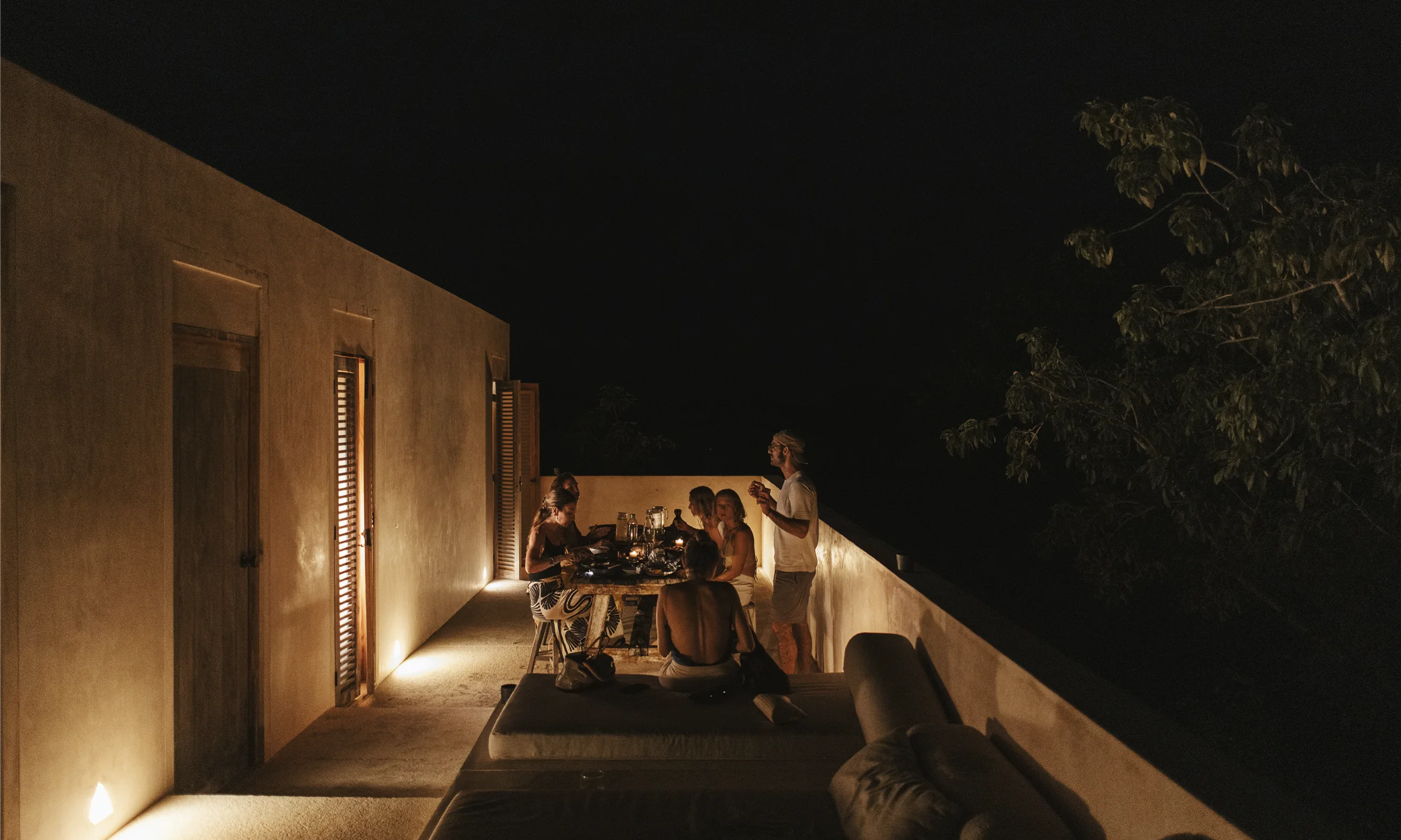 Group of people having dinner and conversation on a dimly lit outdoor terrace at night.