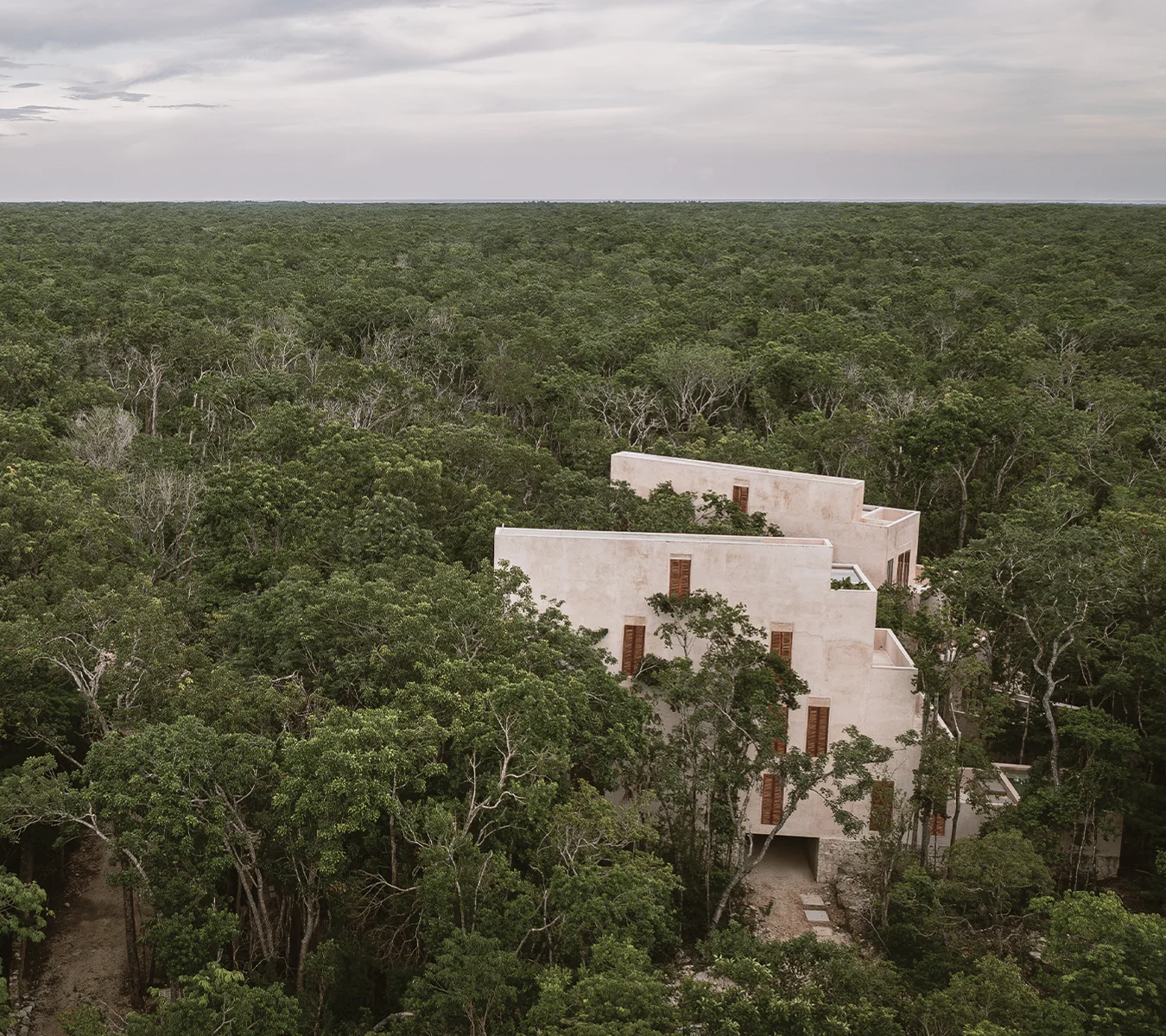 A modern beige building with wooden shutters surrounded by dense green forest under a cloudy sky.