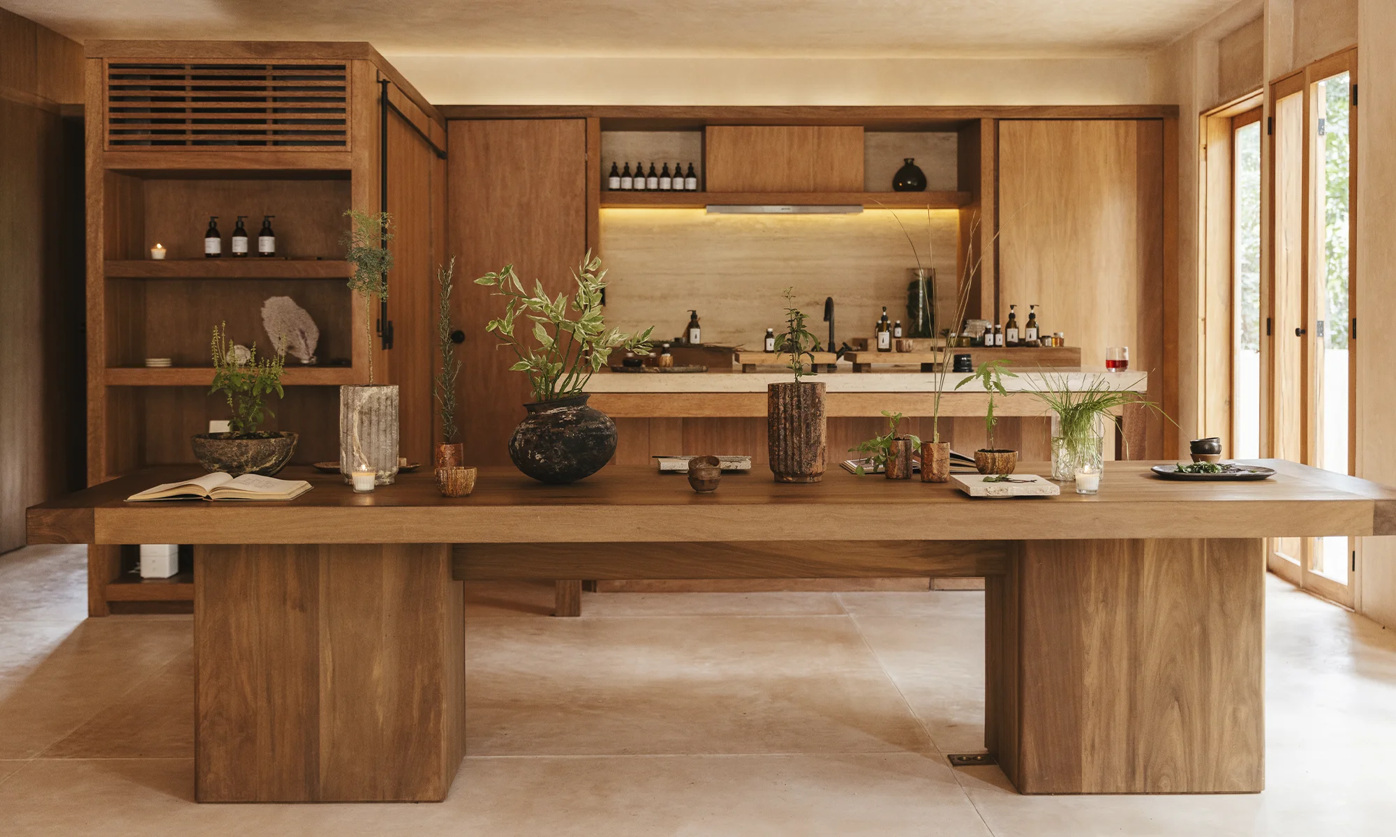 Minimalist wooden kitchen with a large table displaying various potted plants, candles, a book, and glassware, with natural light from large windows.