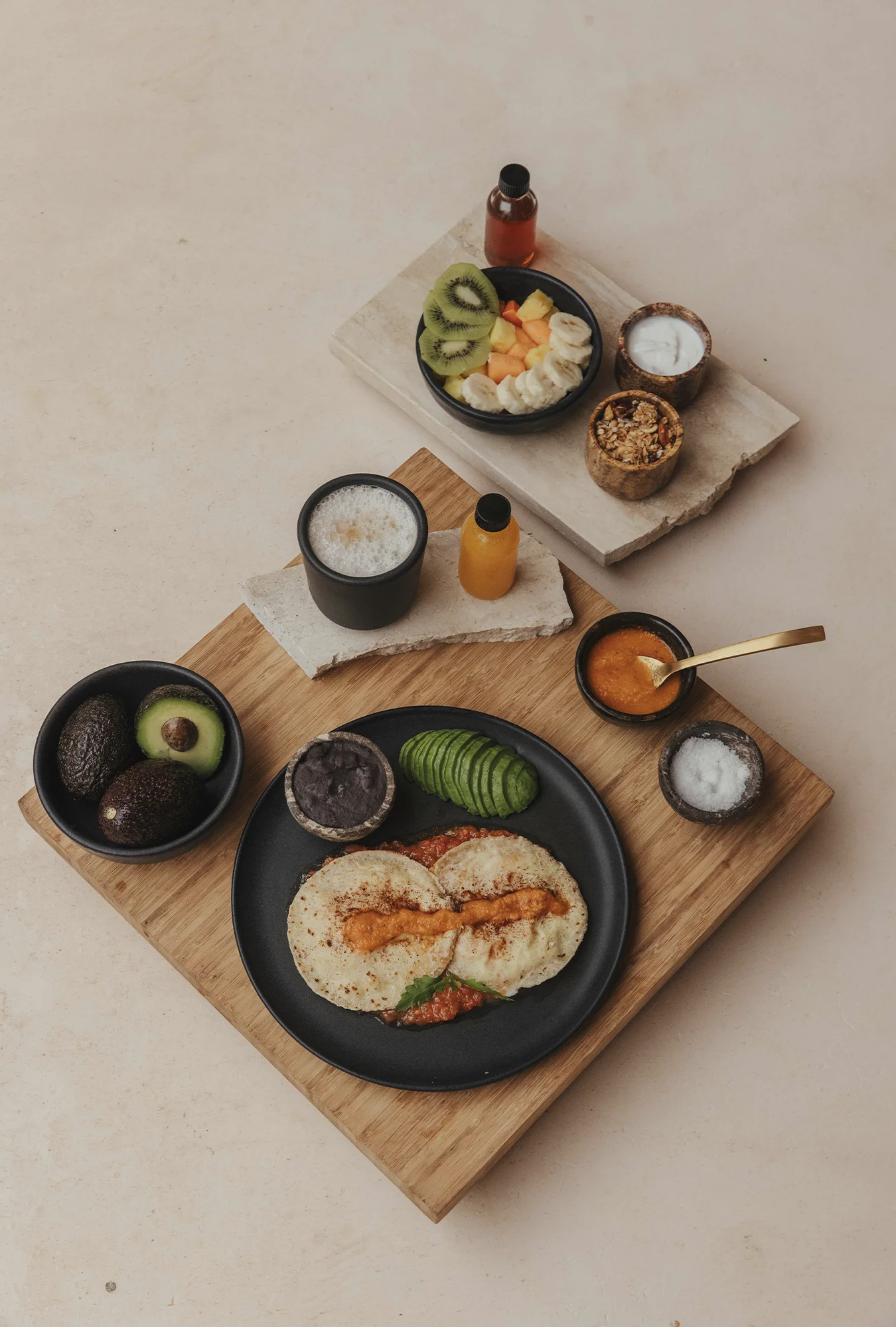 Breakfast spread with two stuffed tortillas on a black plate, sliced avocado, black beans, sauces, whole and halved avocados in a bowl, a cup of frothy coffee, a small bottle of orange juice, and a bowl of mixed fruit with yogurt and granola on rectangular stone trays.