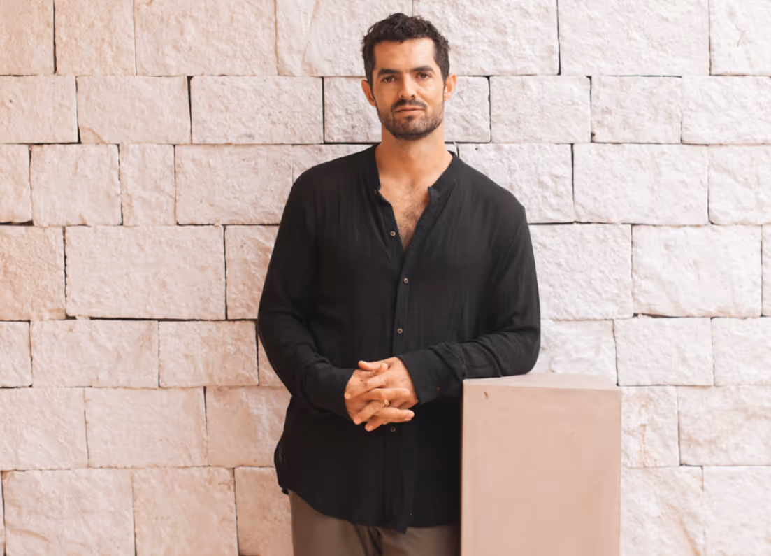 Man with short dark hair and beard wearing a black button-up shirt, standing and leaning on a light-colored pedestal against a textured stone wall.