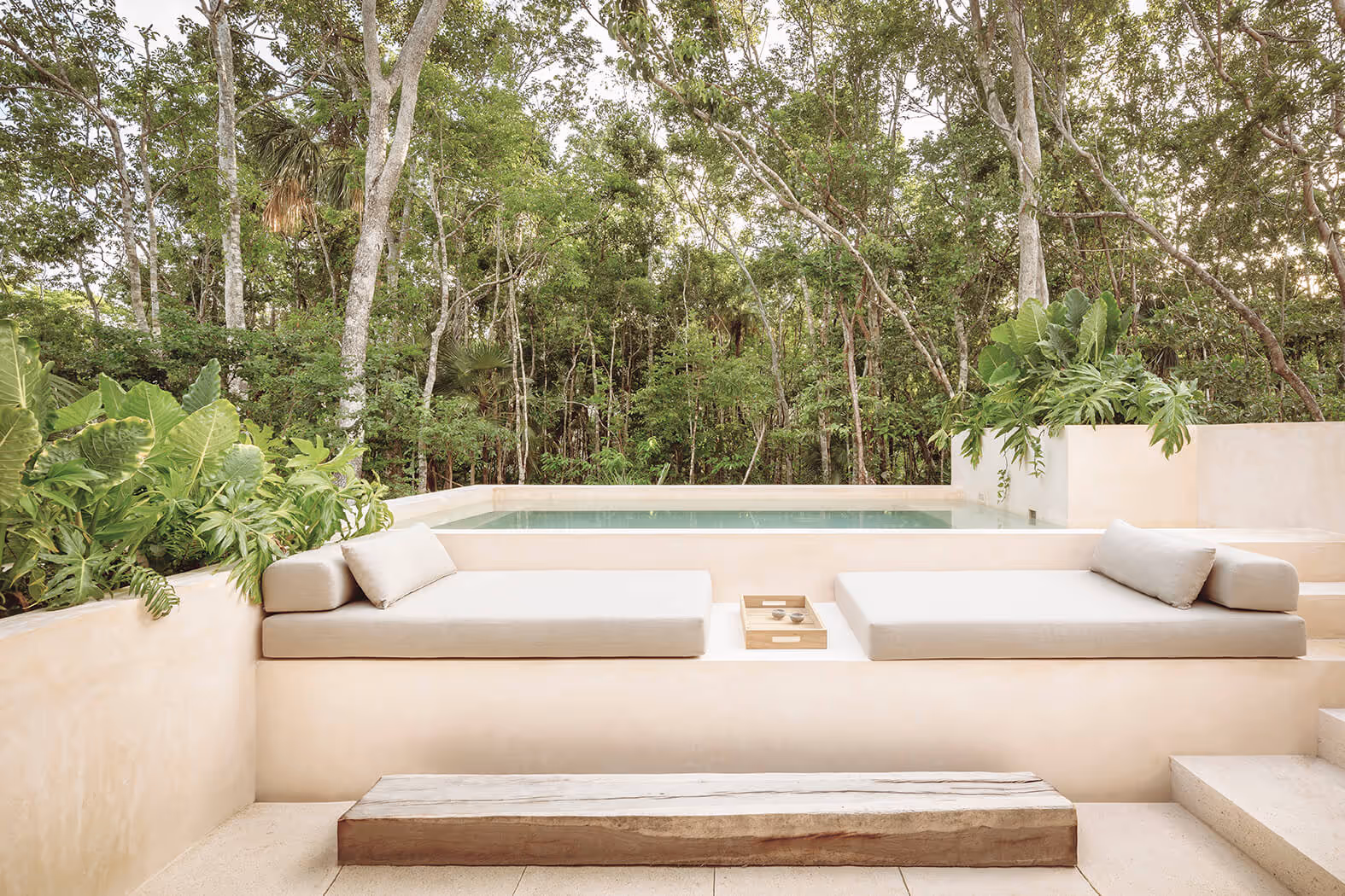 Minimalist outdoor lounge area with beige cushioned seating, a wooden tray, small pool, and lush green forest background.