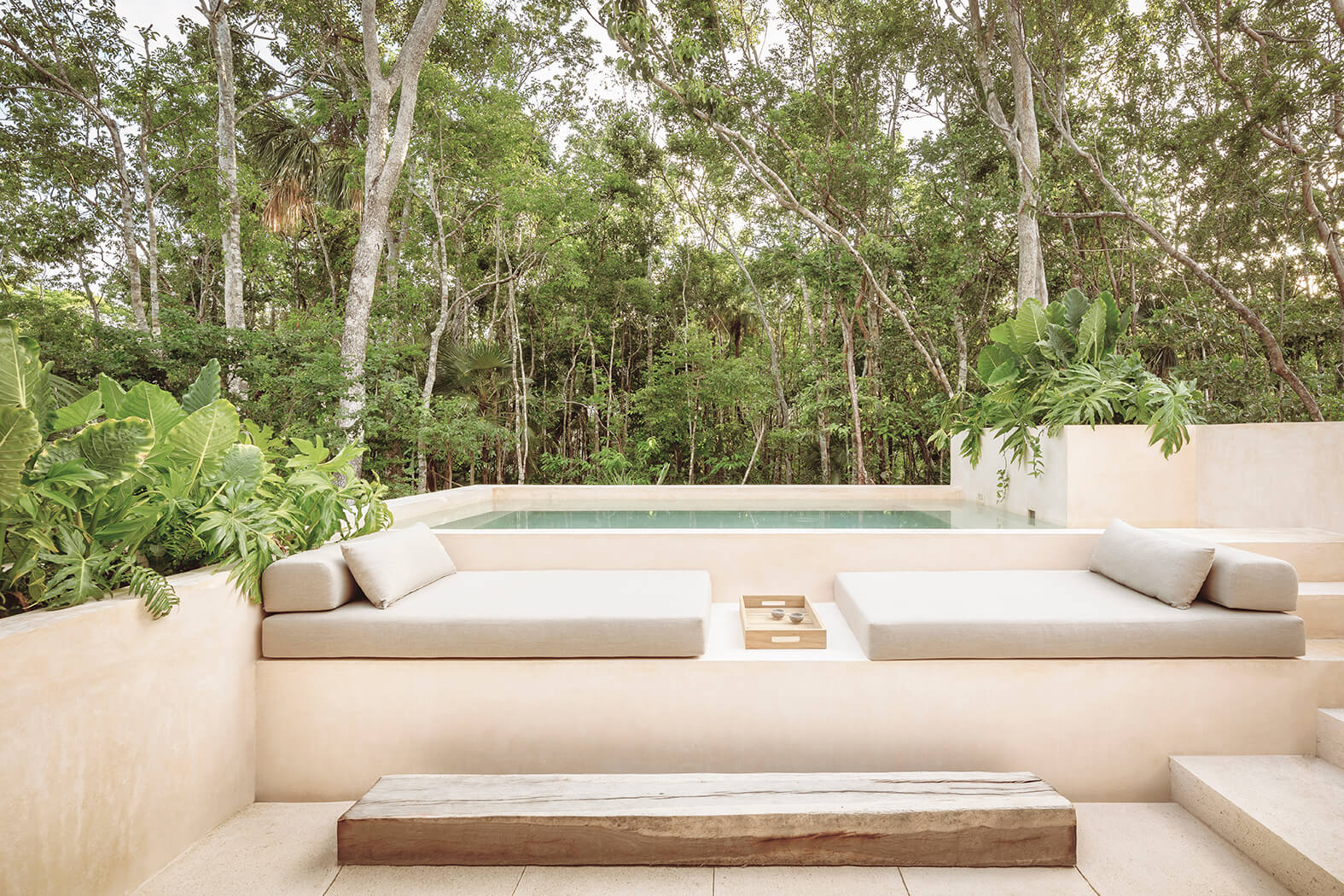 Minimalist outdoor lounge area with beige cushioned seating, a wooden tray, small pool, and lush green forest background.