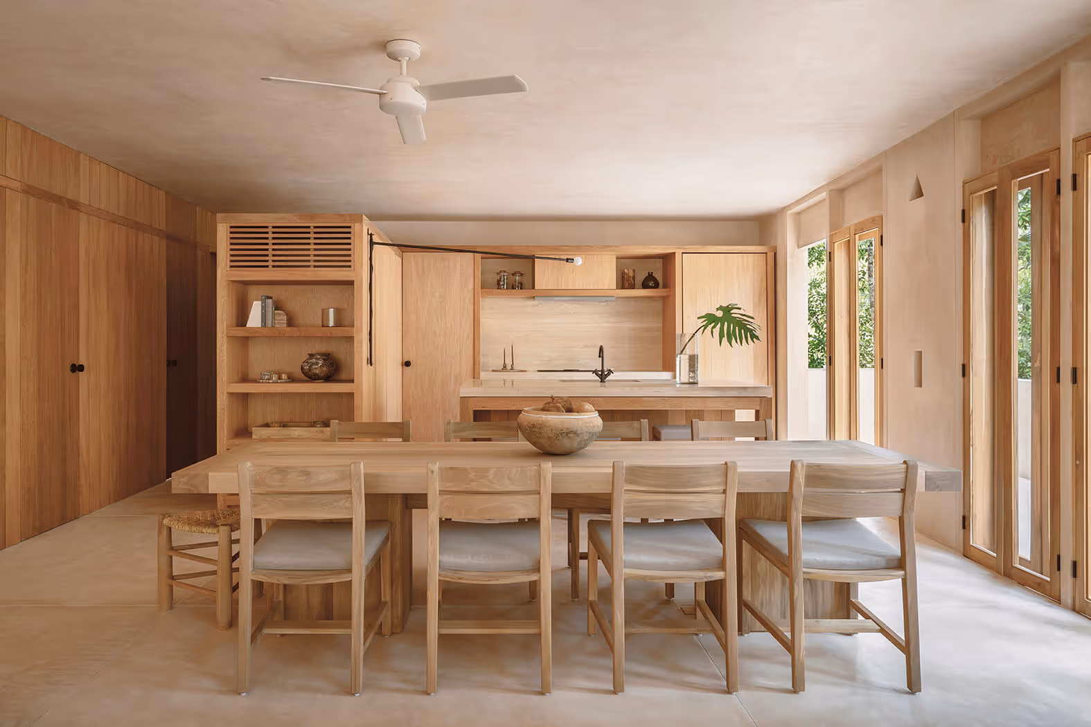 Modern open kitchen and dining area with light wood cabinets, a large wooden dining table with chairs, and glass doors letting in natural light.