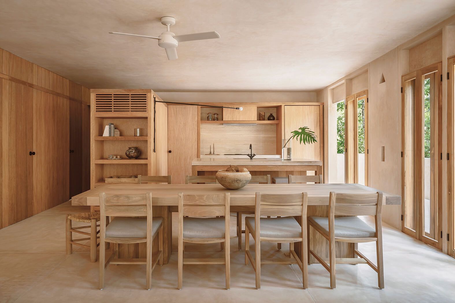 Modern open kitchen and dining area with light wood cabinets, a large wooden dining table with chairs, and glass doors letting in natural light.