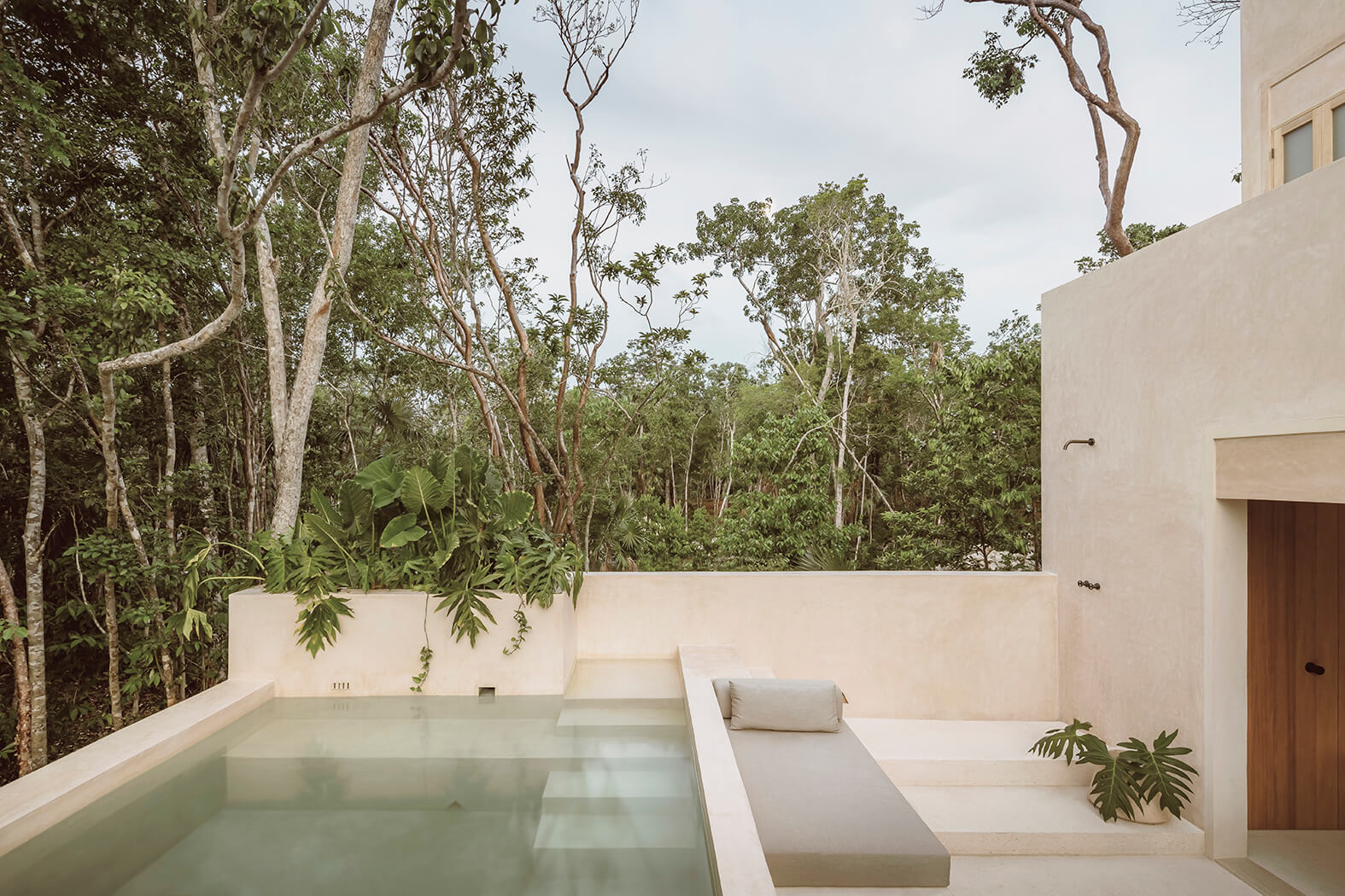 Modern outdoor pool area with beige walls and built-in seating surrounded by lush green forest.