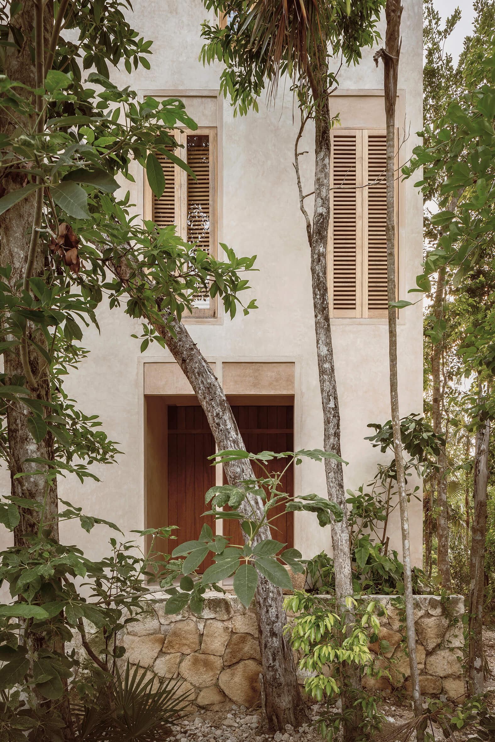 Two-story beige building with wooden doors and shutters surrounded by trees and tropical vegetation.