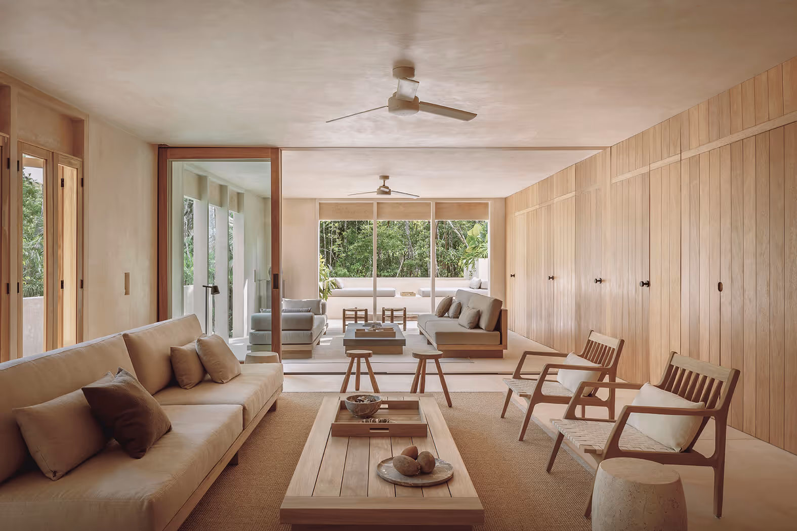Modern spacious living room with beige sofas, wooden chairs, coffee table, and large glass sliding doors showing outdoor greenery.