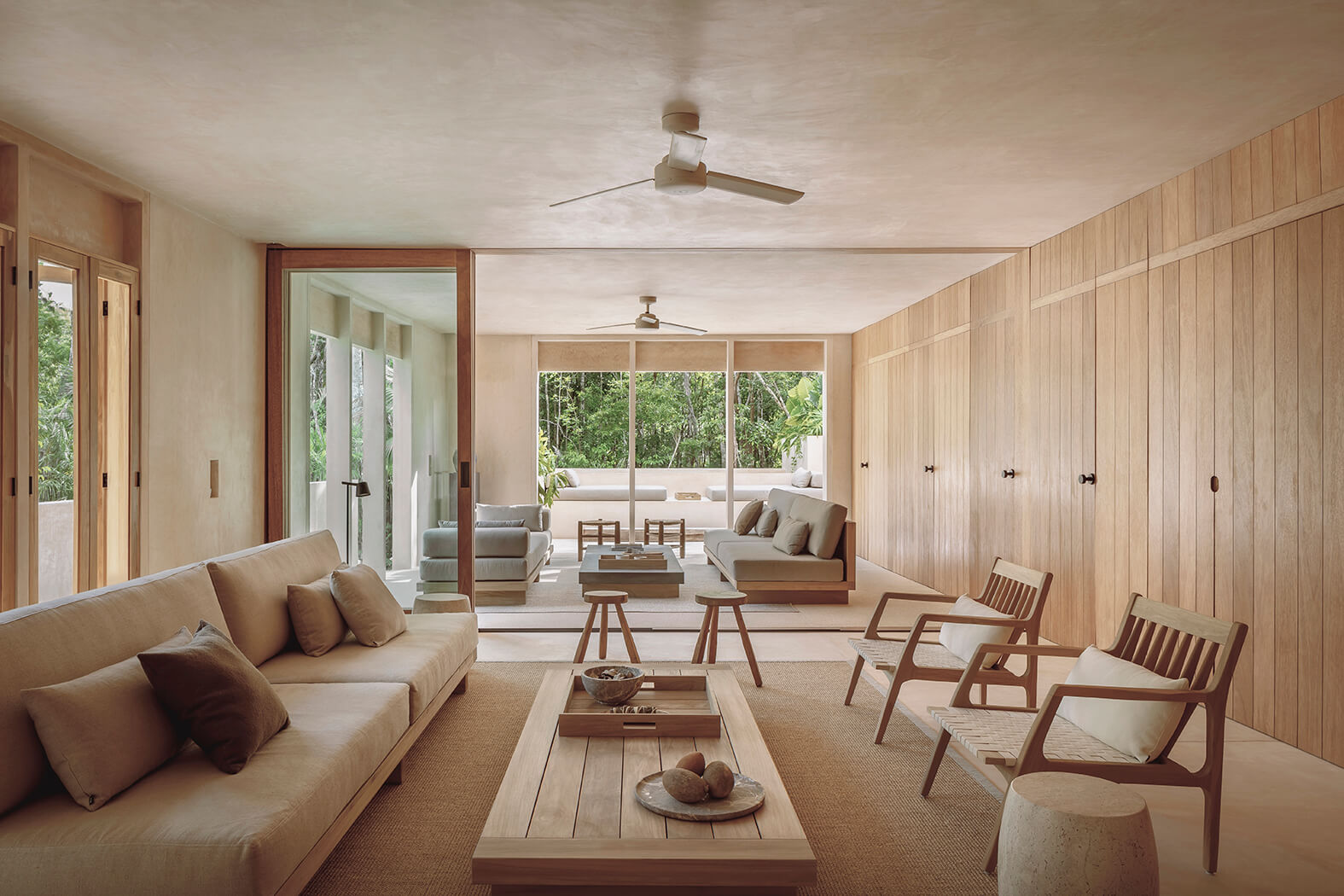 Modern spacious living room with beige sofas, wooden chairs, coffee table, and large glass sliding doors showing outdoor greenery.