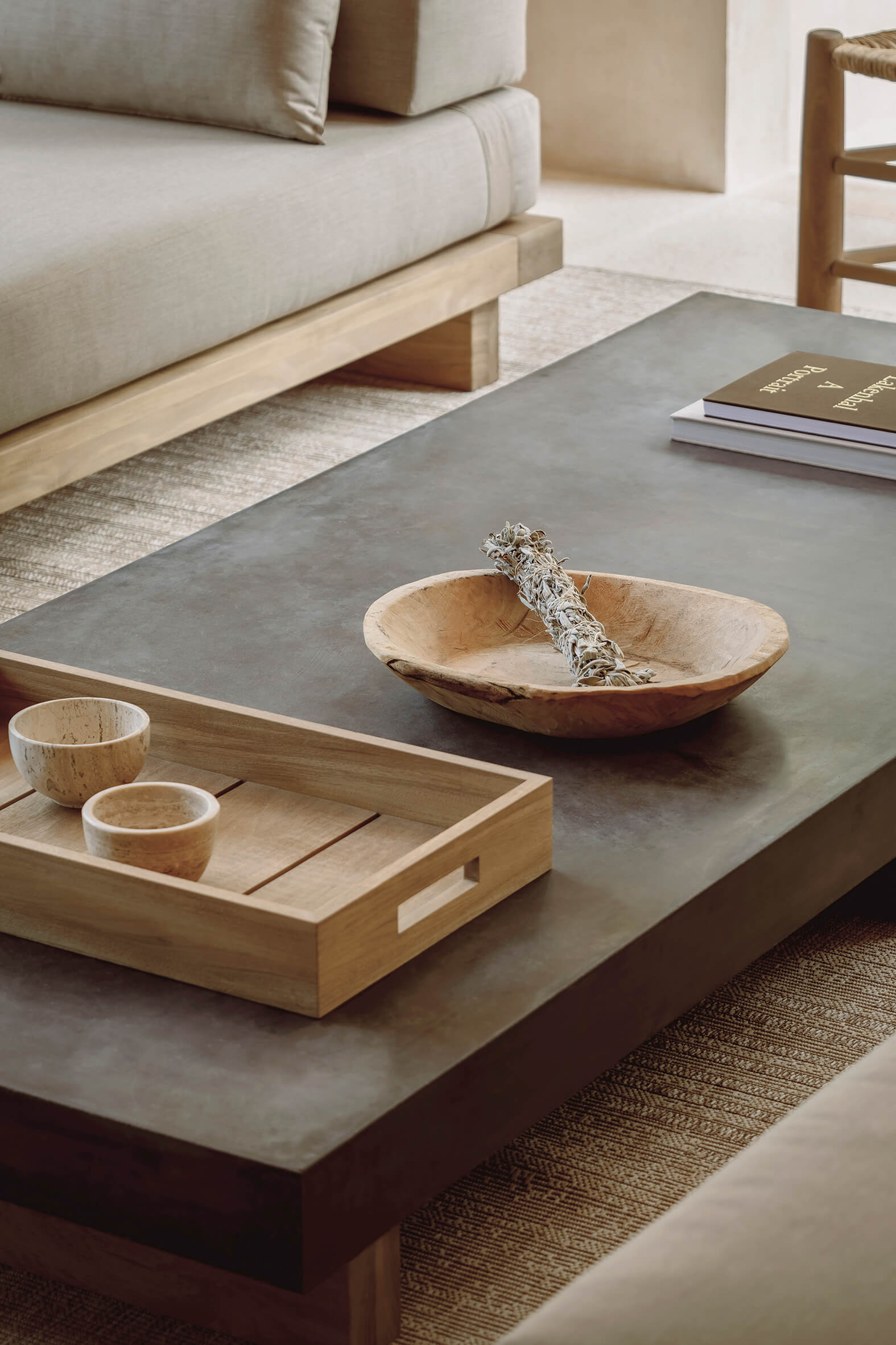 Minimalist living room with a gray sofa, a dark rectangular coffee table topped with a wooden tray holding two small bowls, a wooden dish with a bundle of dried sage, and a stack of books.
