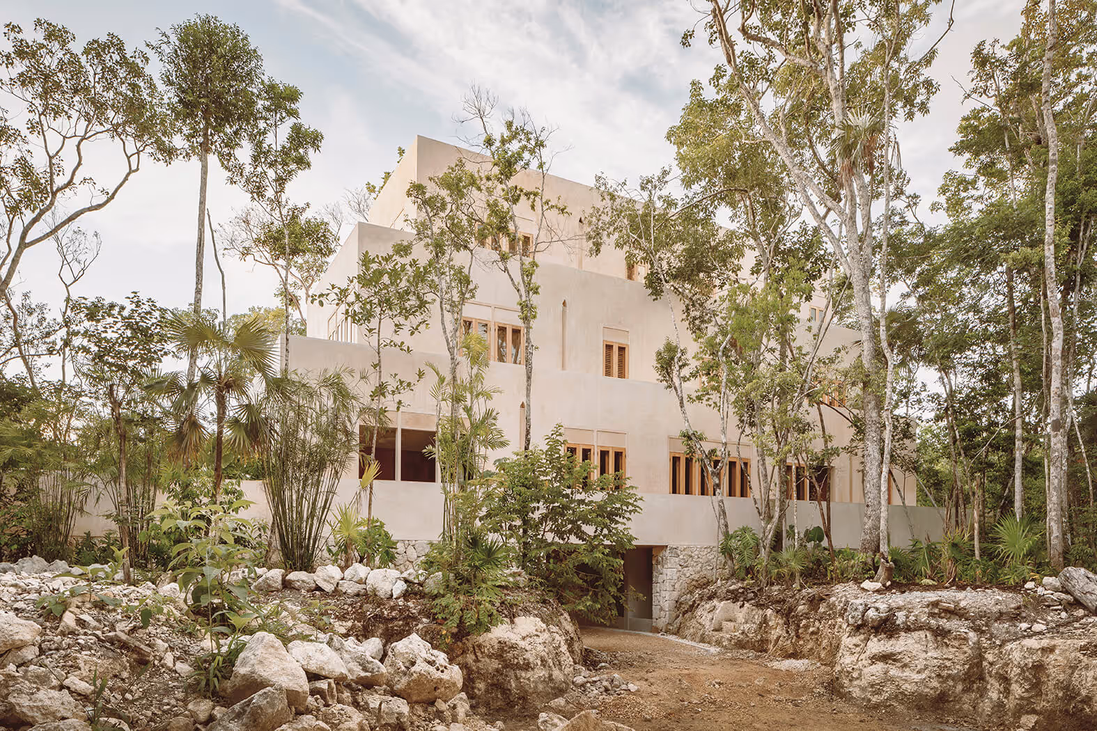 Modern beige multi-story building surrounded by trees and rocky soil under a cloudy sky.