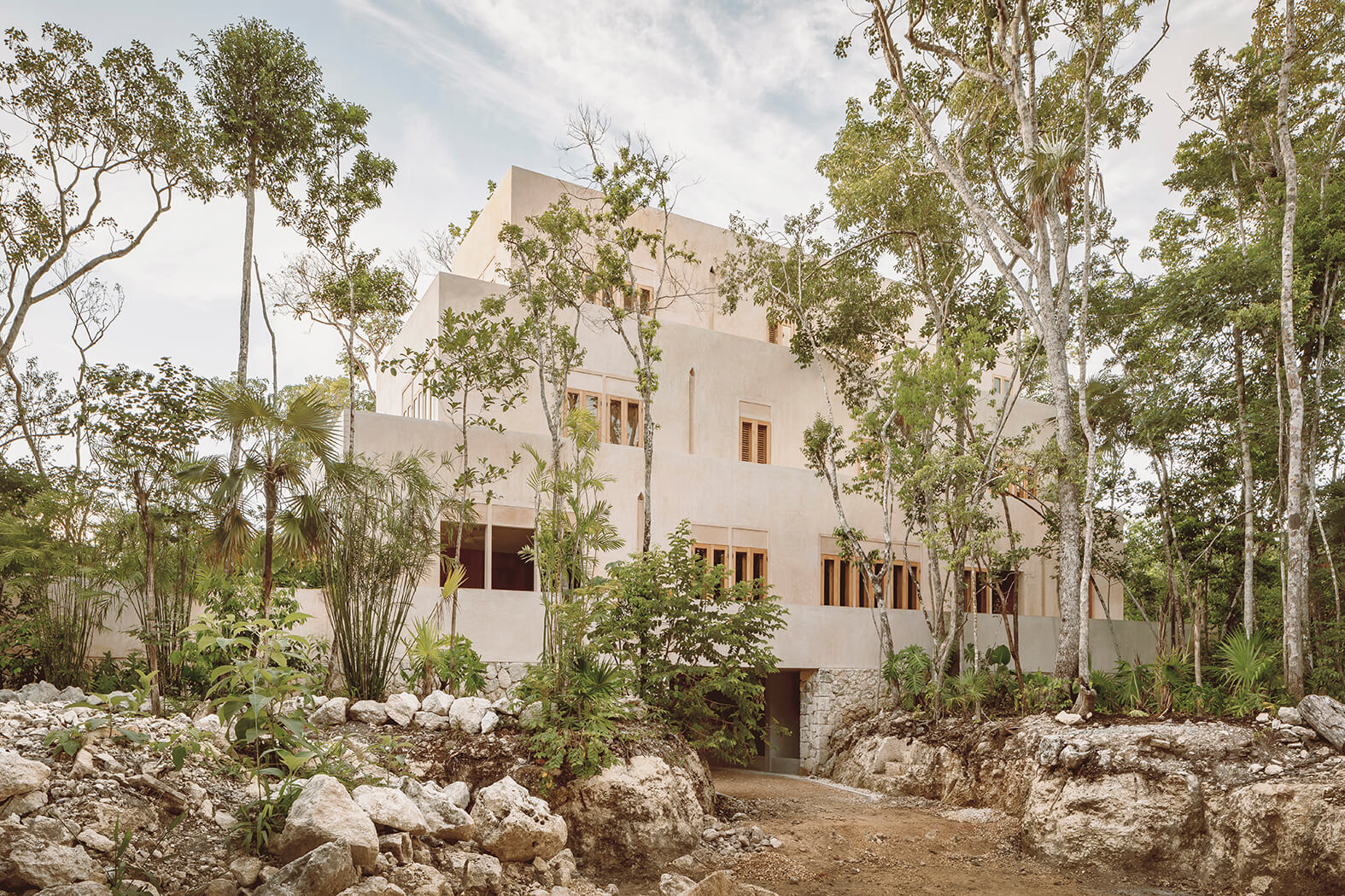 Modern beige multi-story building surrounded by trees and rocky soil under a cloudy sky.