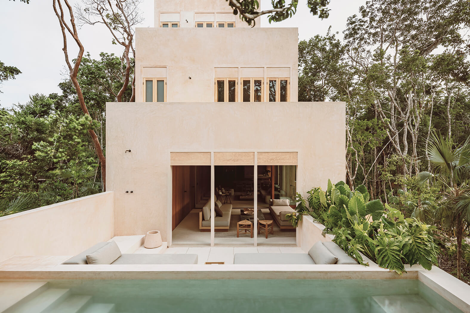 Modern beige stucco house with large glass doors opening to a seating area by a pool surrounded by lush green trees.