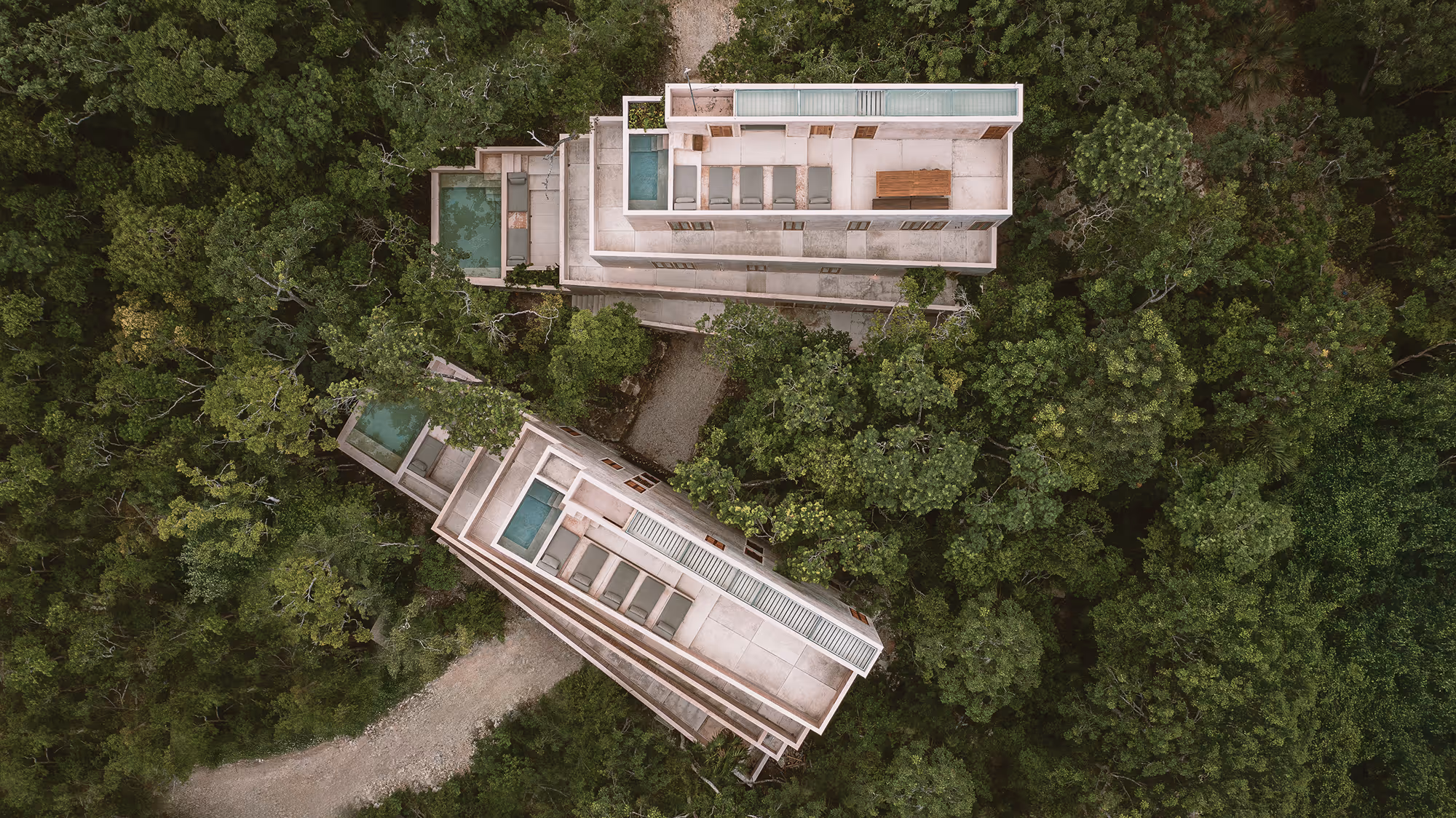 Aerial view of two modern villas with pools surrounded by dense green forest and a dirt path.