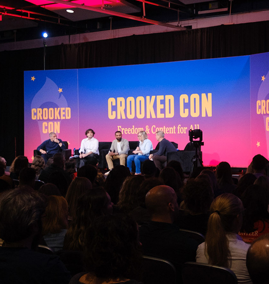 Panelists sit on stage on front of large CROOKED CON backdrop.