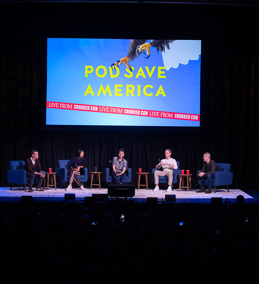 Panelists sit on stage with Pod Save America graphic projected above them.