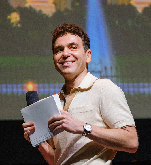 Jon Lovett smiles holding microphone and papers.