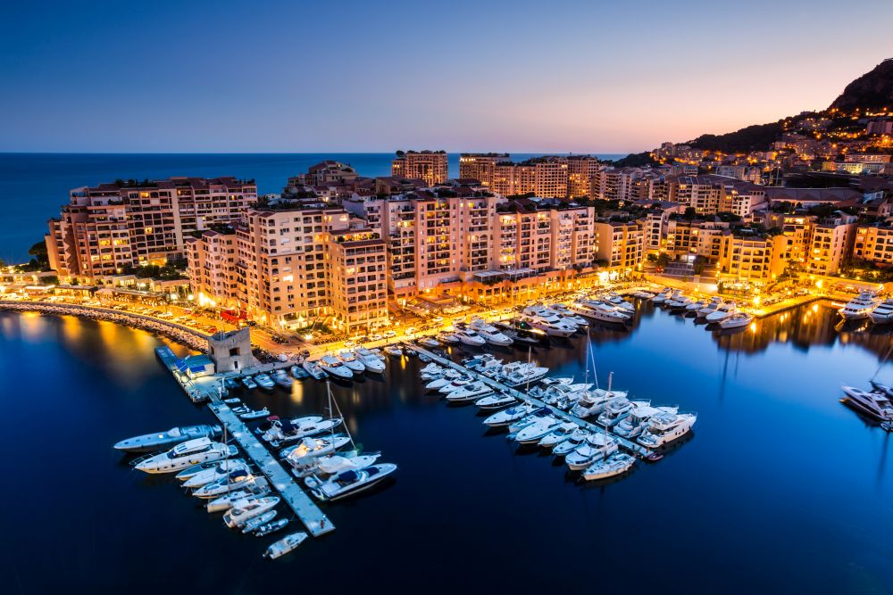 A panoramic view of Monte Carlo harbor featuring luxury yachts, the Monaco skyline, and Mediterranean Sea with mountains in the background.