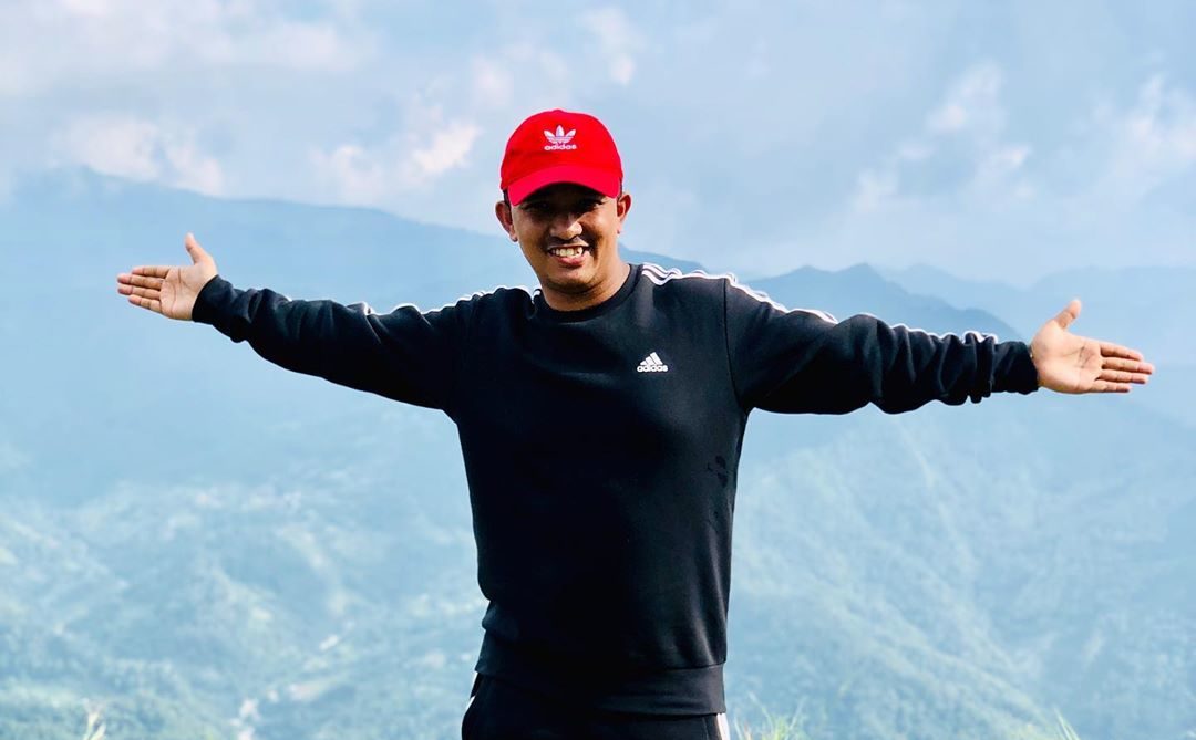 Carnival Cruise Line crew member Safiq Ahmed stands with his arms outstretched in front of a mountain range. He sports a red baseball cap and black Adidas pullover. Safiq relies on Brightwell's global payroll solution, Navigator, for access to his personal finances. He sends remittances back home to India to help support his family.
