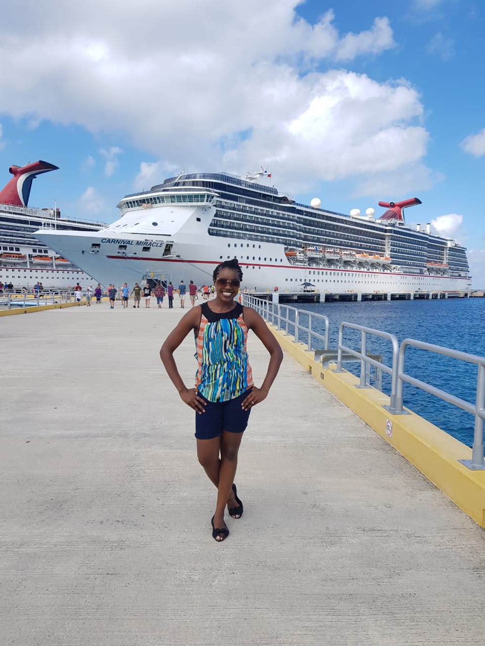 Carnival Cruise Line crew member Perpetual Kanyemba smiles with her hands on her hips in front of the Carnival Miracle ship. She receieves her pay via Brightwell's global payroll solution, Navigator, which aims to empower global workers by providing quick, secure, and efficient access to finances.