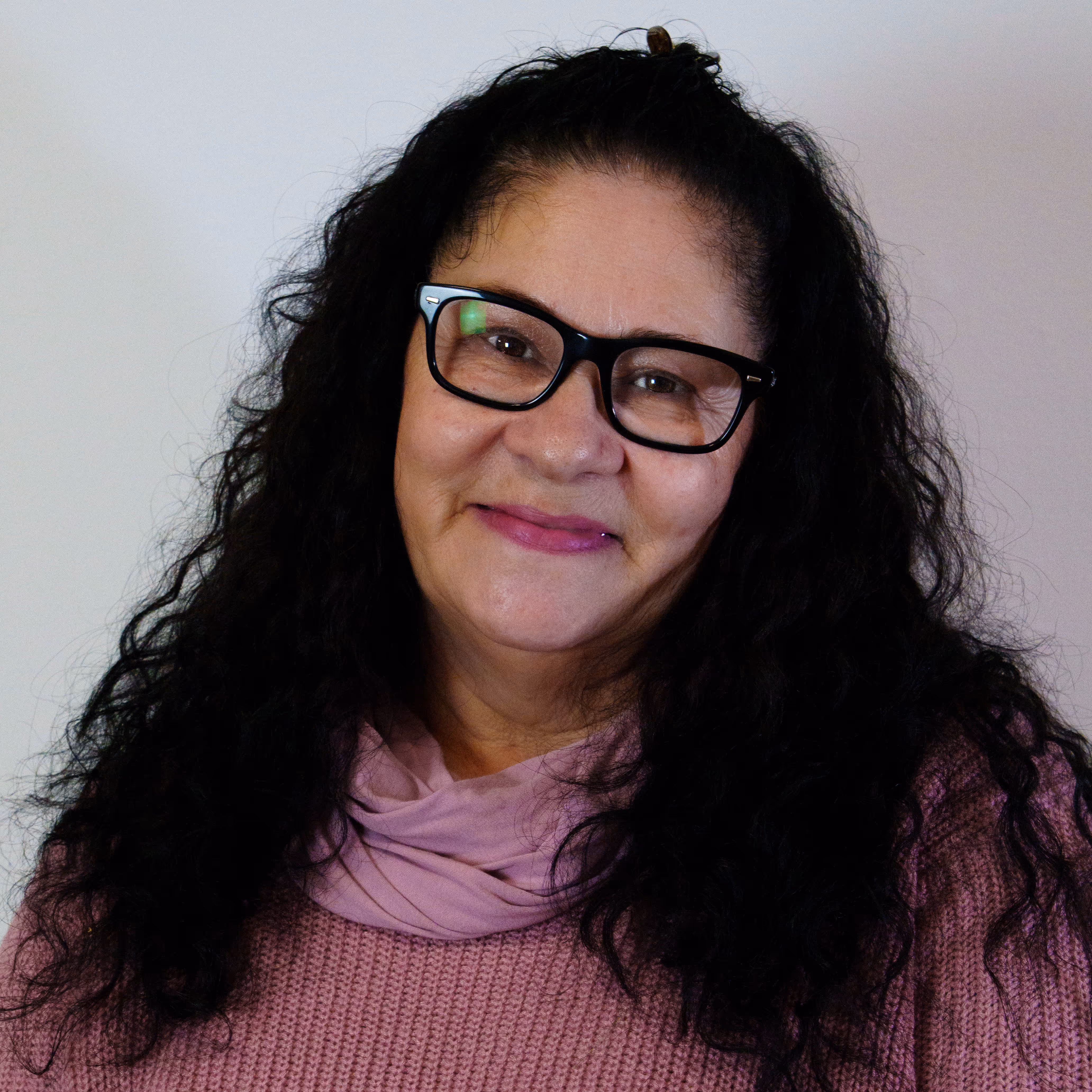 Smiling woman with long curly black hair, black glasses, and a pink sweater and scarf.