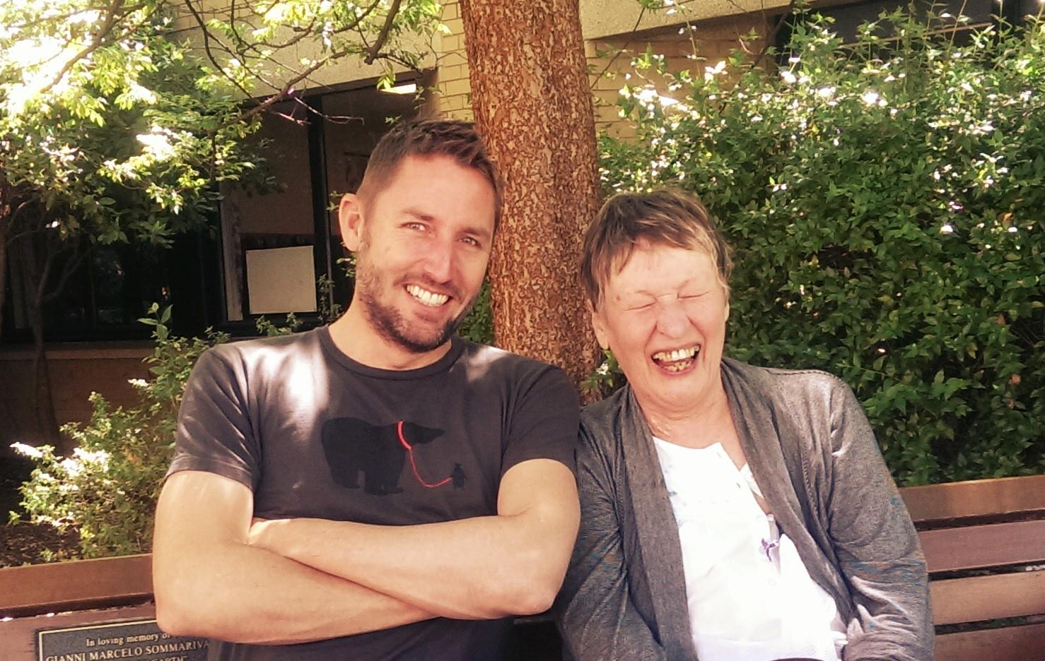 Andrew Ferguson and Elizabeth Mamchak sitting together on a bench outdoors, with trees and greenery in the background.
