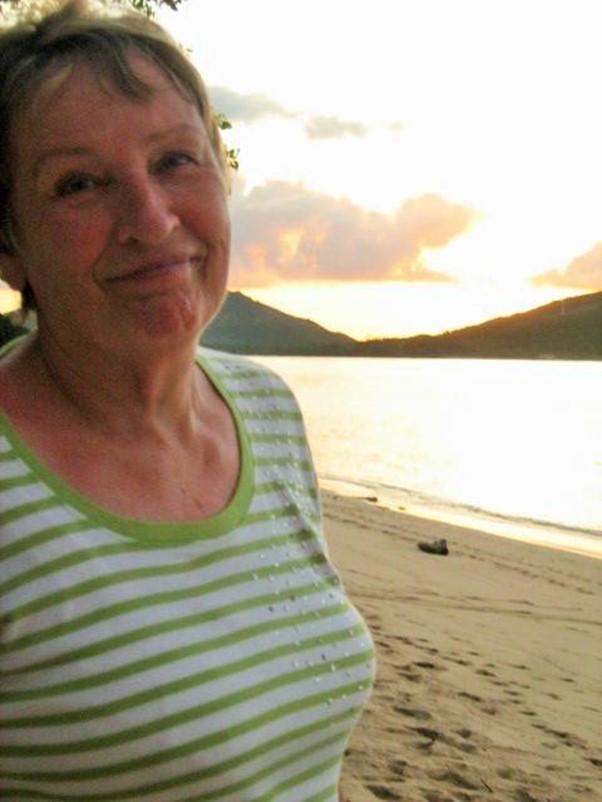 Elizabeth Mamchak in Fiji, smiling in a green and white striped shirt standing on a sandy beach at sunset with mountains in the background.
