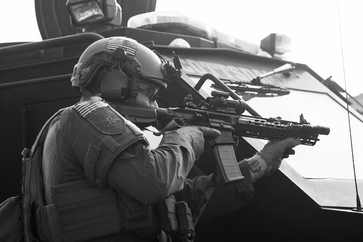 SWAT officer in tactical gear aiming a rifle beside an armored police vehicle.