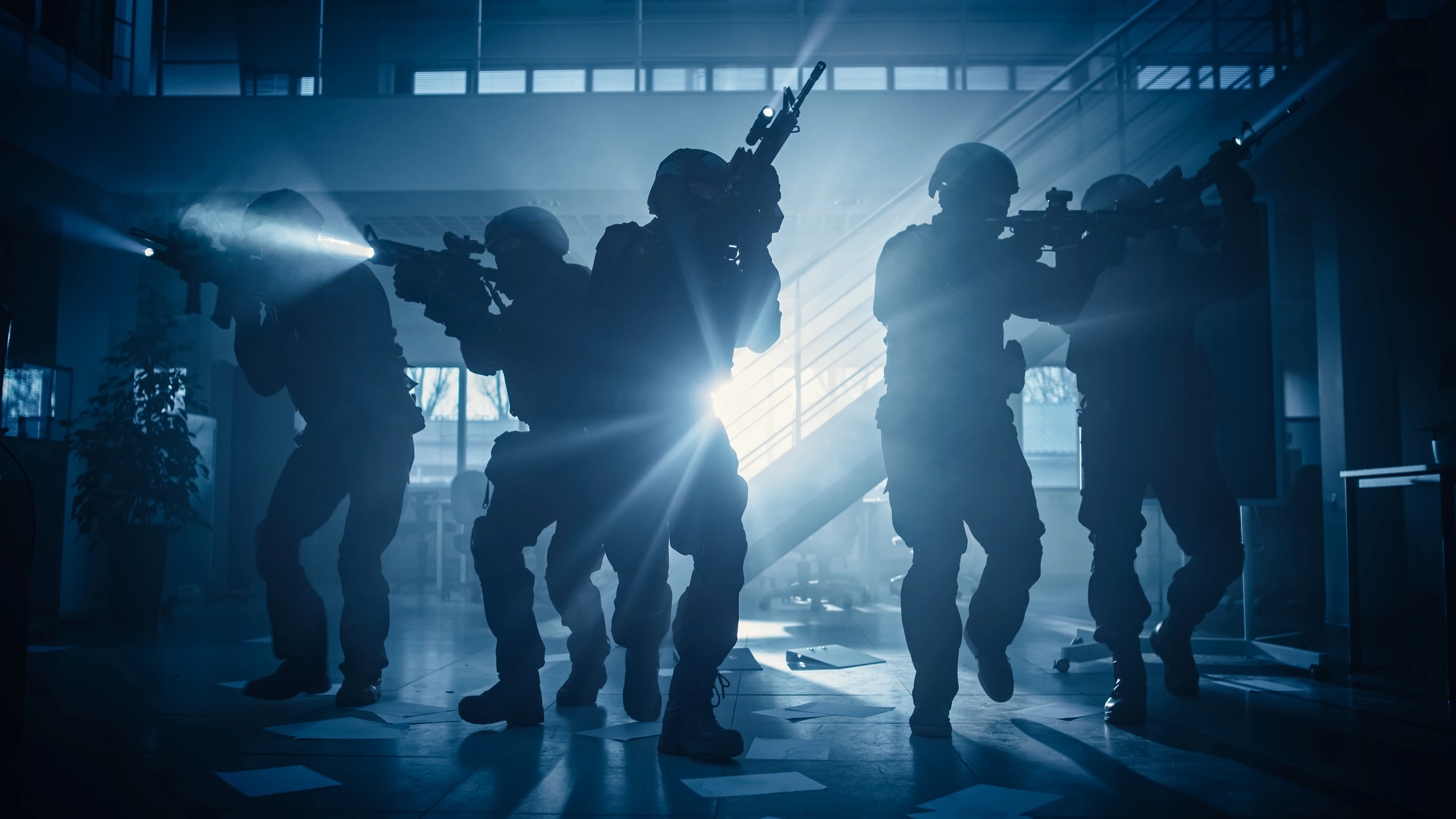 Silhouettes of tactical officers with rifles and flashlights advancing inside a dimly lit building.