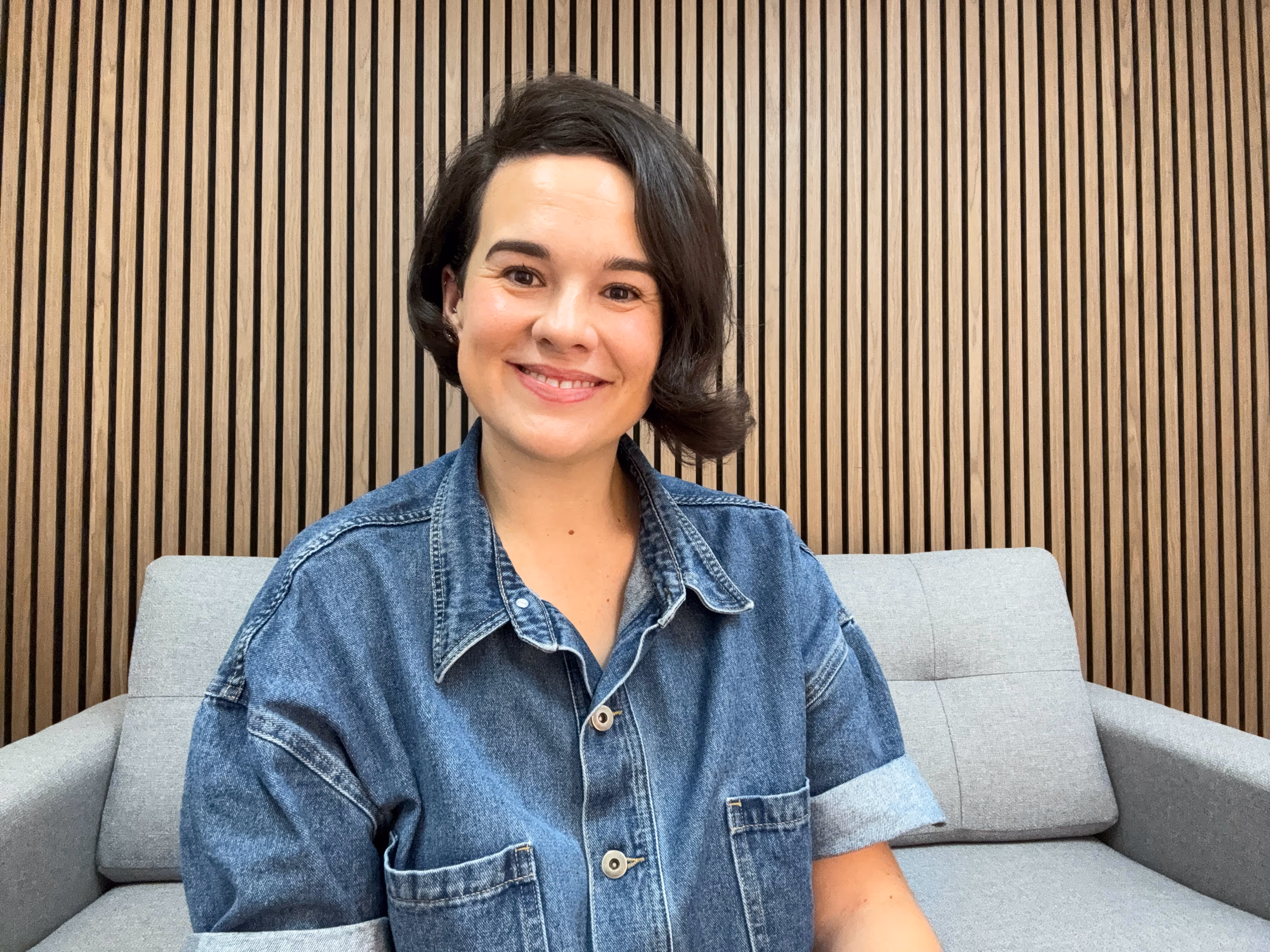 Smiling woman with short dark hair wearing a denim shirt sitting on a gray couch with a wooden slat wall background.