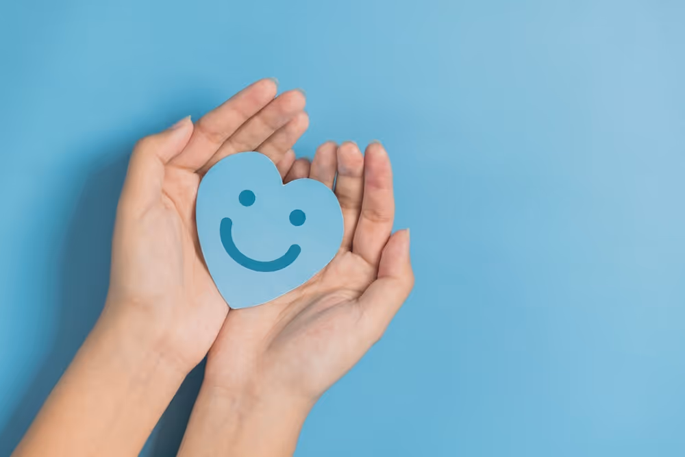 Hands gently holding a blue paper heart with a smiling face on a matching blue background.