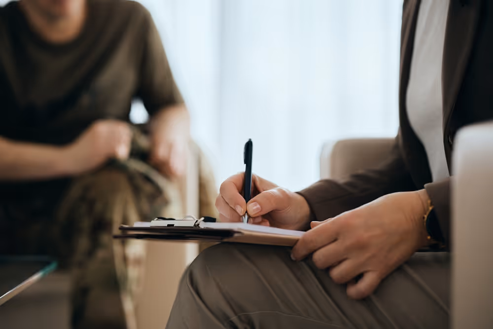 Person writing notes on a clipboard during a conversation with someone in military attire.