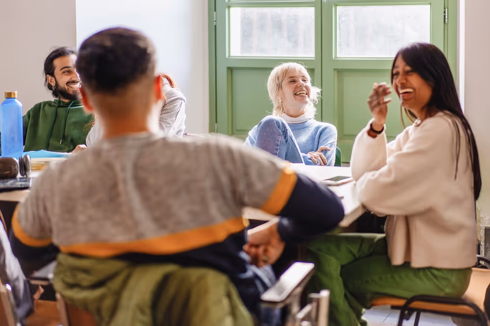 A group of five young adults sitting around a table, smiling and laughing together in a bright room with green window frames.
