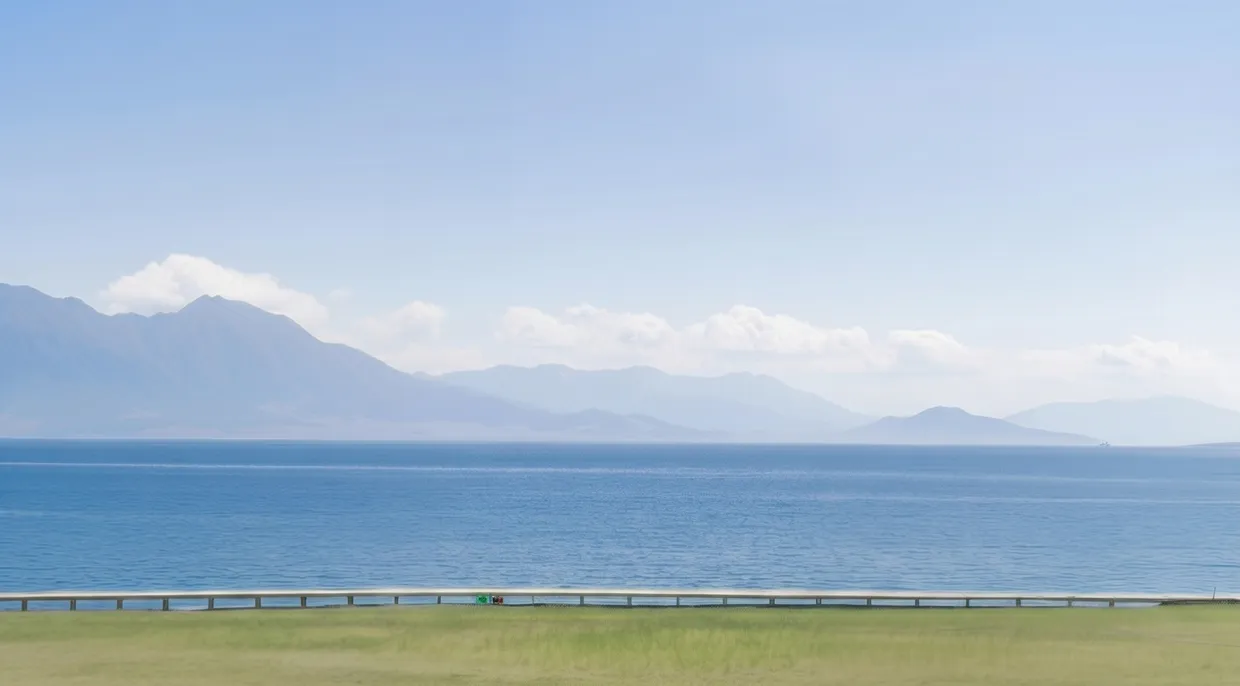 Calm blue sea with distant mountain range under a clear sky and a green grassy foreground.