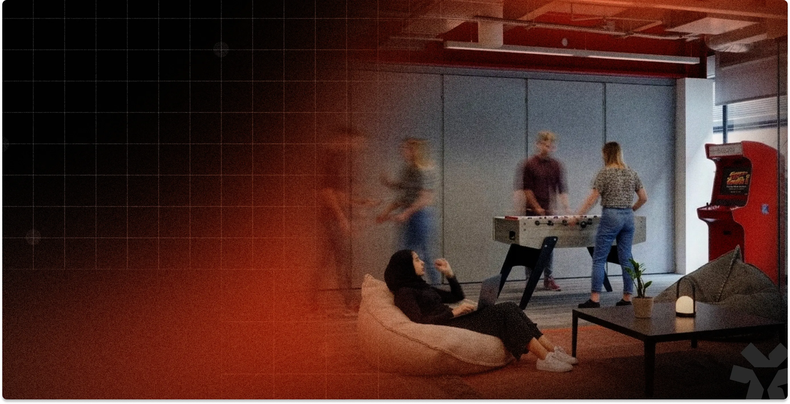 Casual office space with one person sitting on a beanbag using a laptop, and three people playing foosball near a red arcade machine.