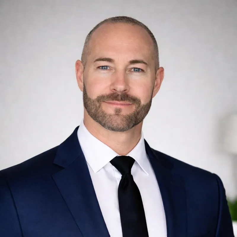 Professional man with light skin, blue eyes, and a beard wearing a navy suit, white shirt, and black tie, smiling slightly in a studio setting.