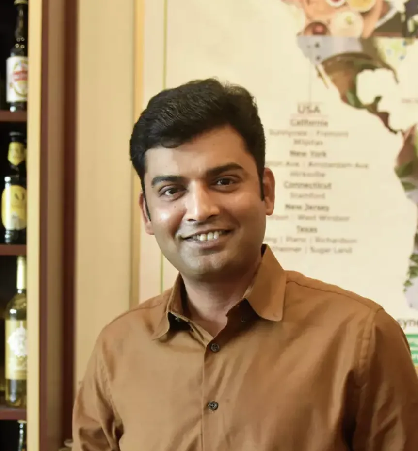 Smiling man with short black hair wearing a brown button-up shirt standing indoors with a shelf of bottles and a map behind him.