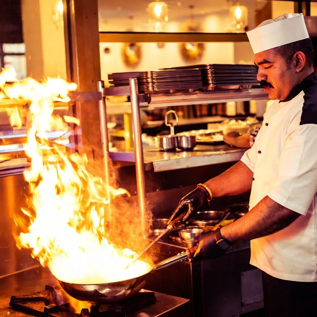 Chef wearing a white hat and black gloves cooking with high flames in a commercial kitchen.