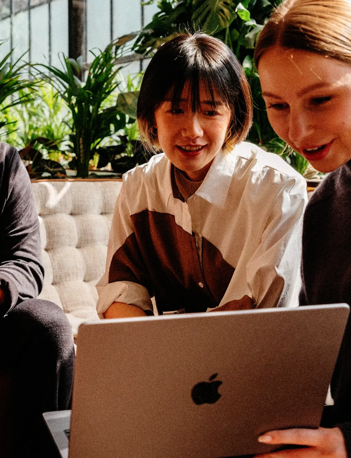 Two women sitting on a sofa looking at a silver MacBook laptop, with green plants in the background.