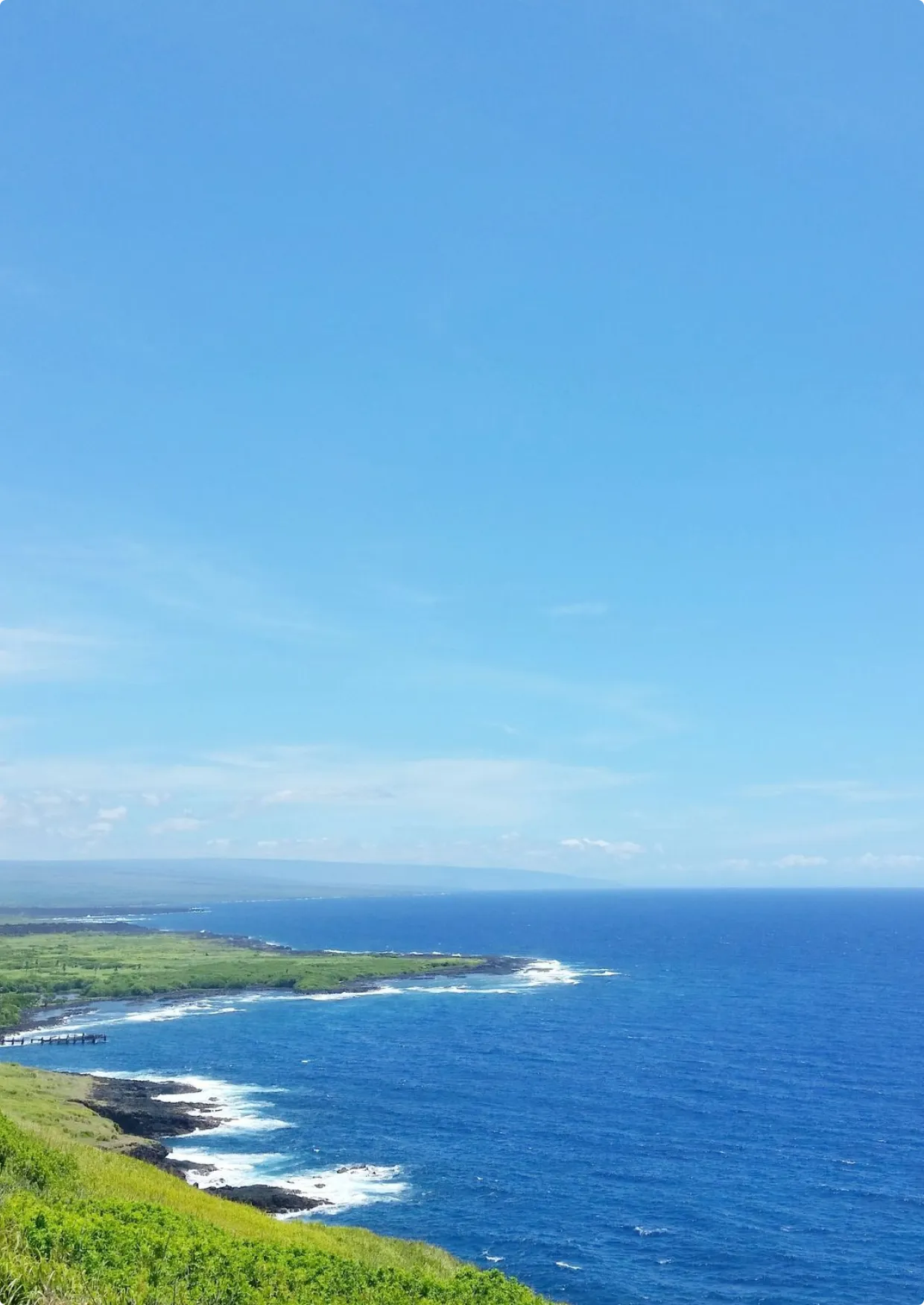 Coastal landscape with green hills, rocky shore, and deep blue ocean under a clear sky.