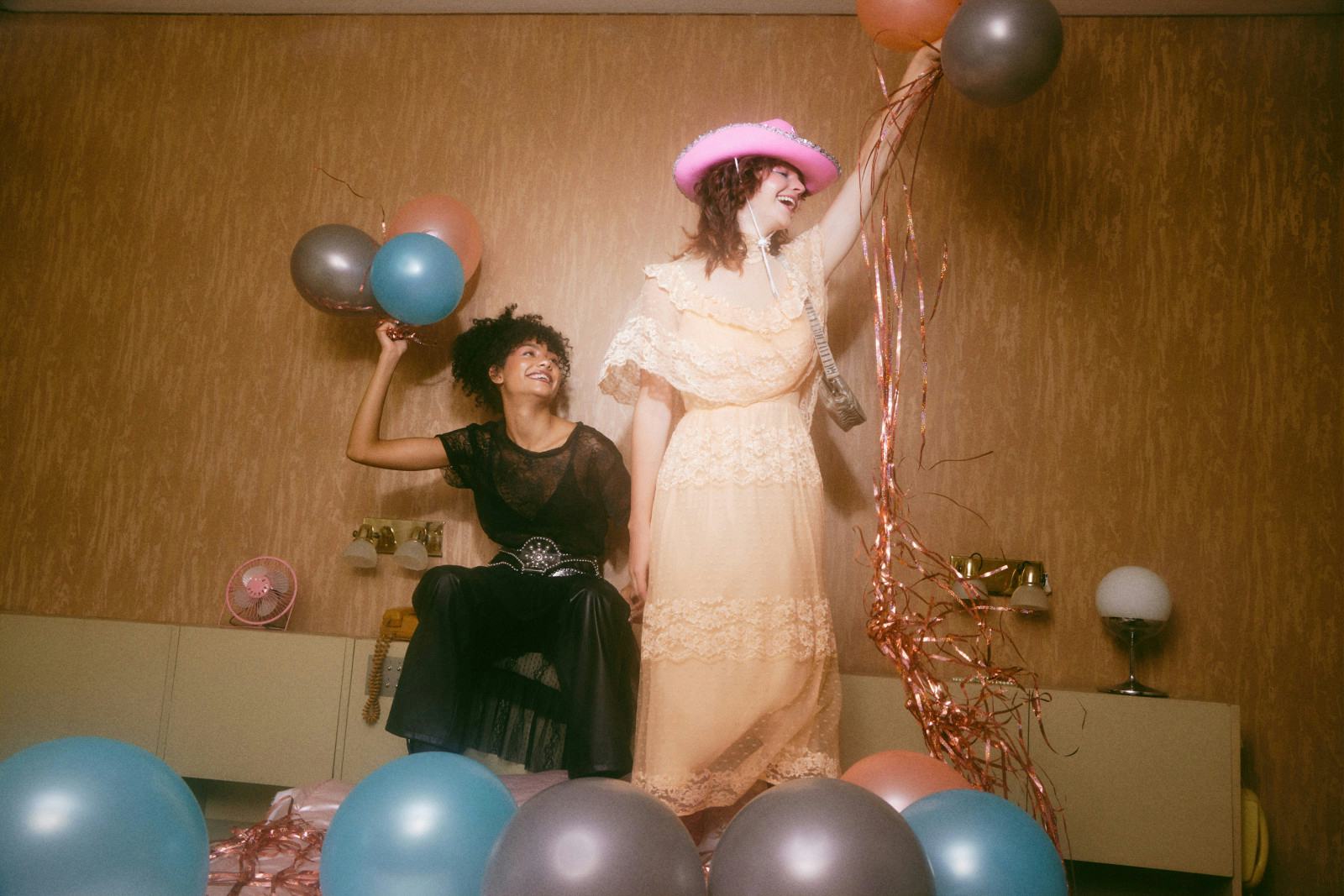 Two joyful young women holding balloons in a room decorated with blue, gray, and pink balloons, one seated and the other standing wearing a pink cowboy hat and peach dress.