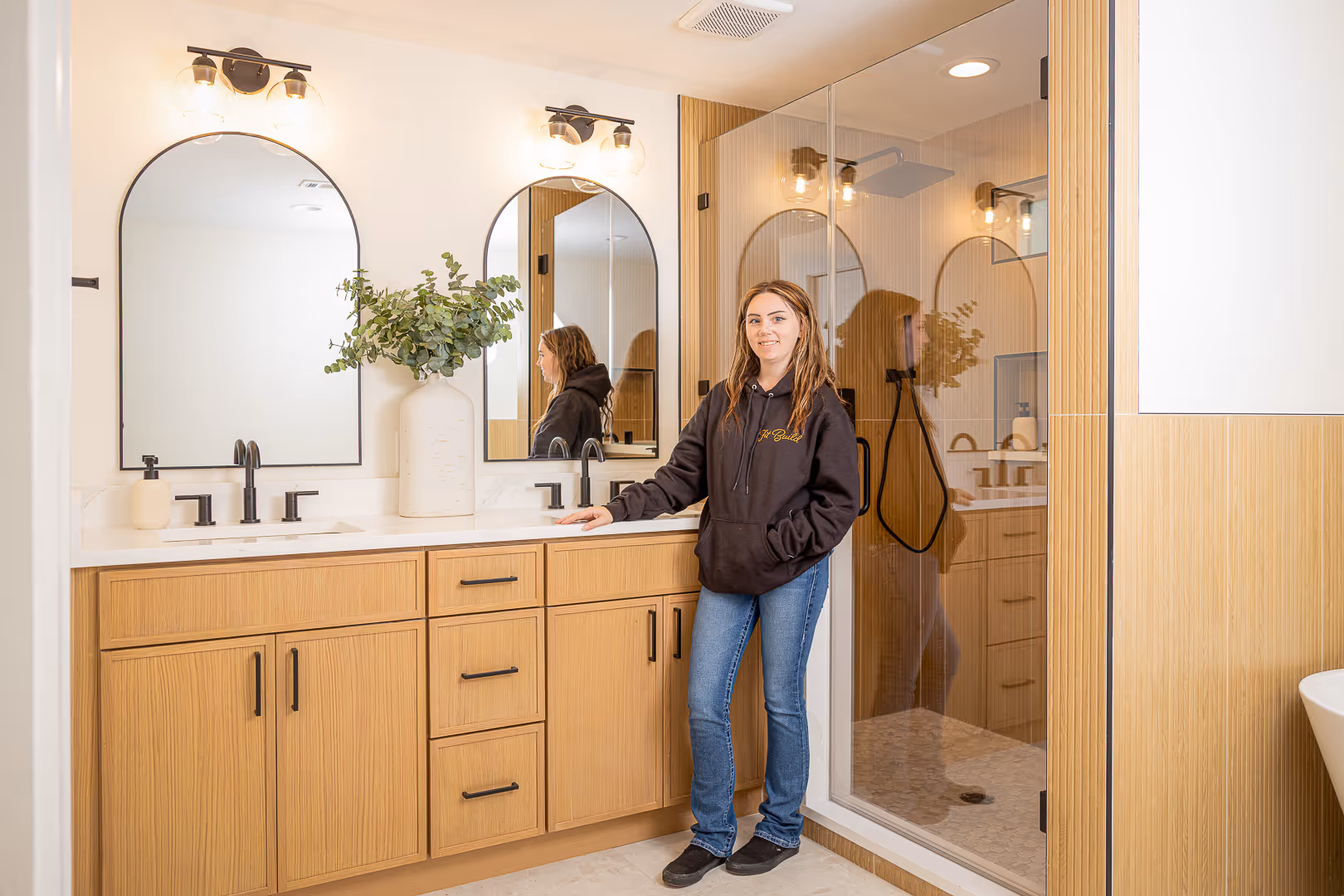 Woman in a black hoodie and jeans standing in a modern bathroom with wooden cabinets, dual mirrors, and a glass shower.