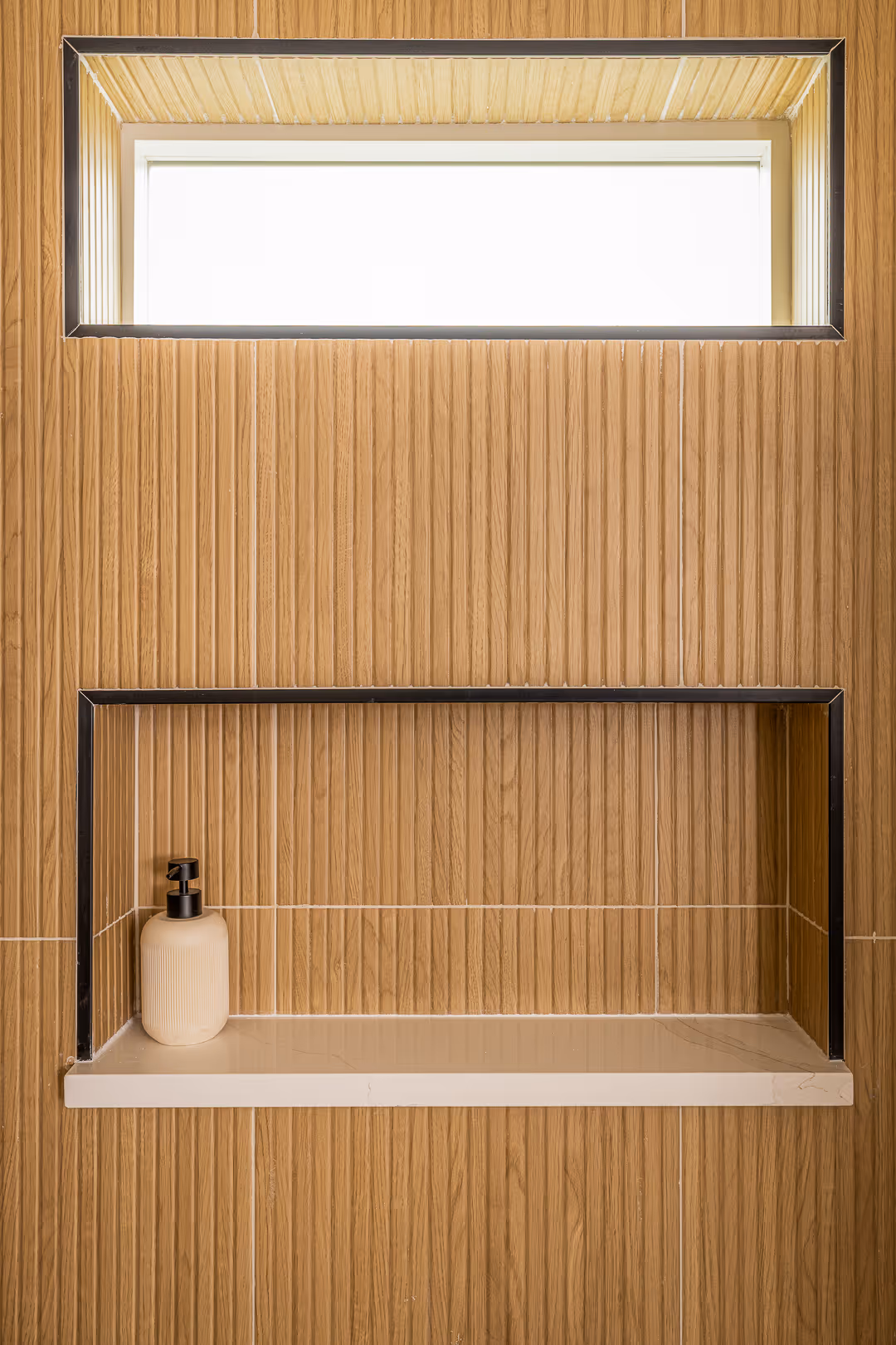 Wall with vertical wooden panels featuring a recessed shelf holding a beige soap dispenser and a rectangular window above it.