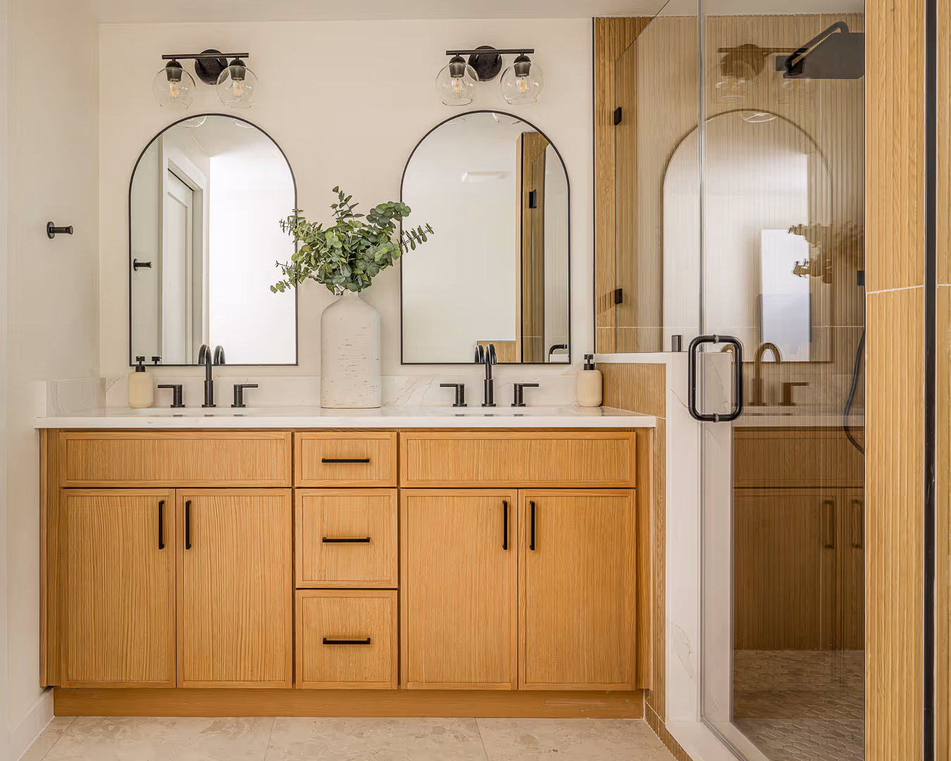 Modern bathroom with double wooden vanity featuring black fixtures, two arched mirrors, a large white vase with greenery, and a glass shower enclosure.